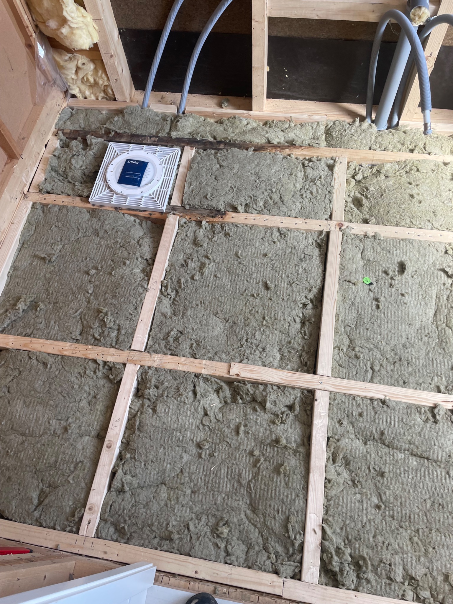 Montaż izolacji z wełny mineralnej między drewnianymi legarami poddasza, widoczne szare rury instalacyjne i biała kratka wentylacyjna