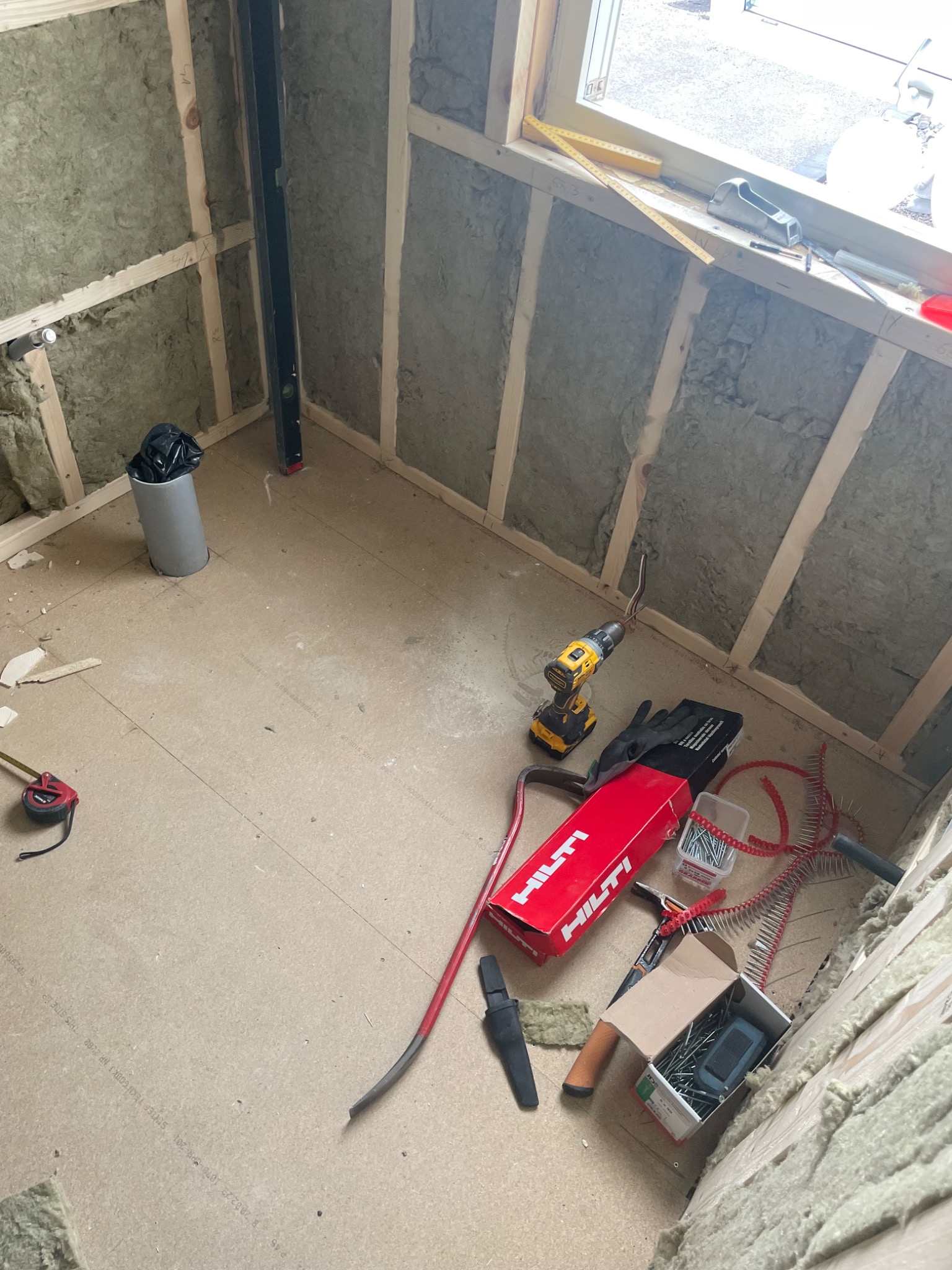 Wnętrze w trakcie ocieplania wełną mineralną, widoczne narzędzia pracy: wiertarka, poziomica, miarka, pudełko gwoździ Hilti, przewody i rury