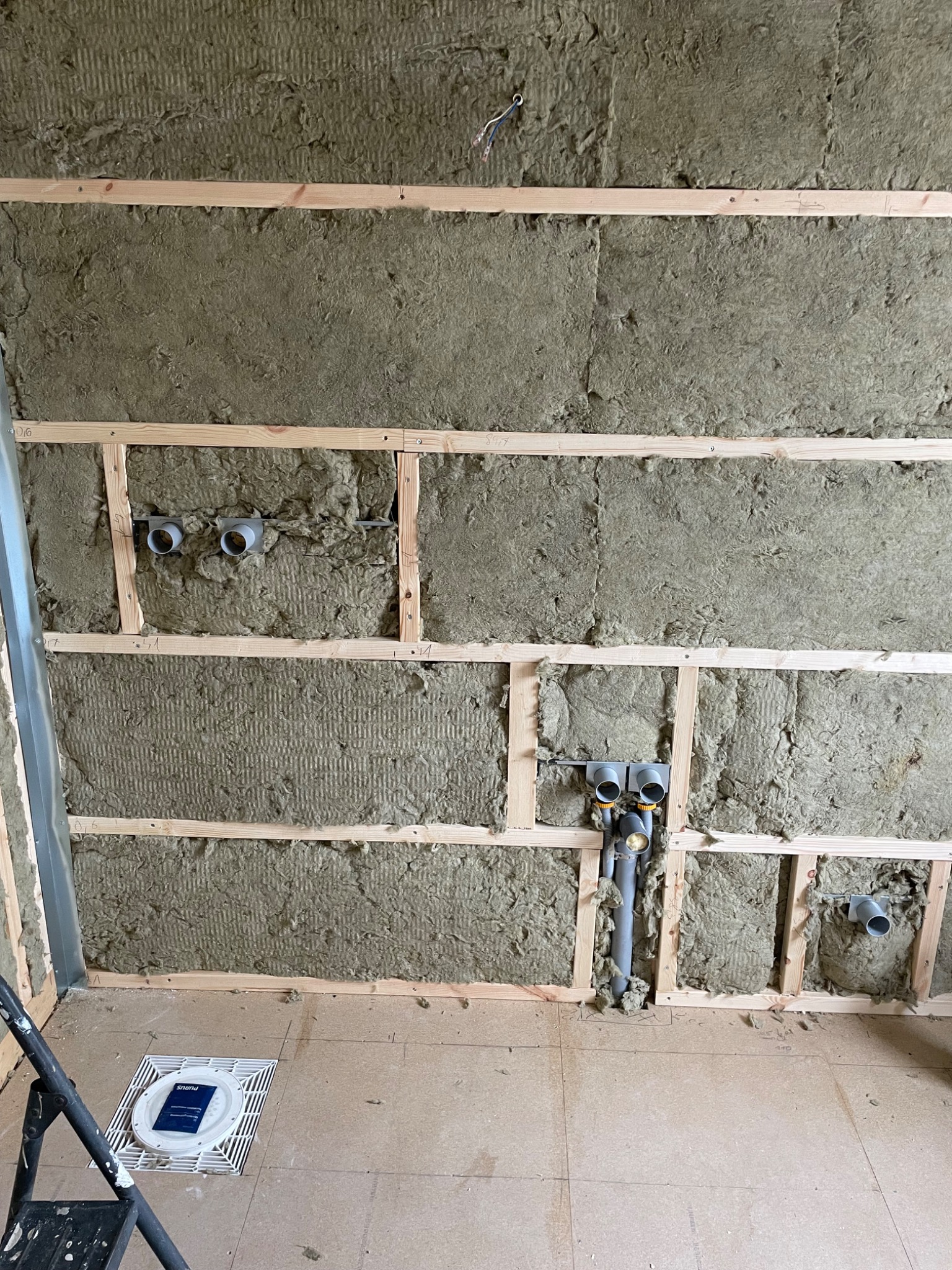 Ściana w trakcie izolacji wełną mineralną, widoczne drewniane ramy, instalacja kanalizacyjna i odpływ podłogowy z białym rusztem, na podłodze płyty OSB z naniesionymi oznaczeniami.