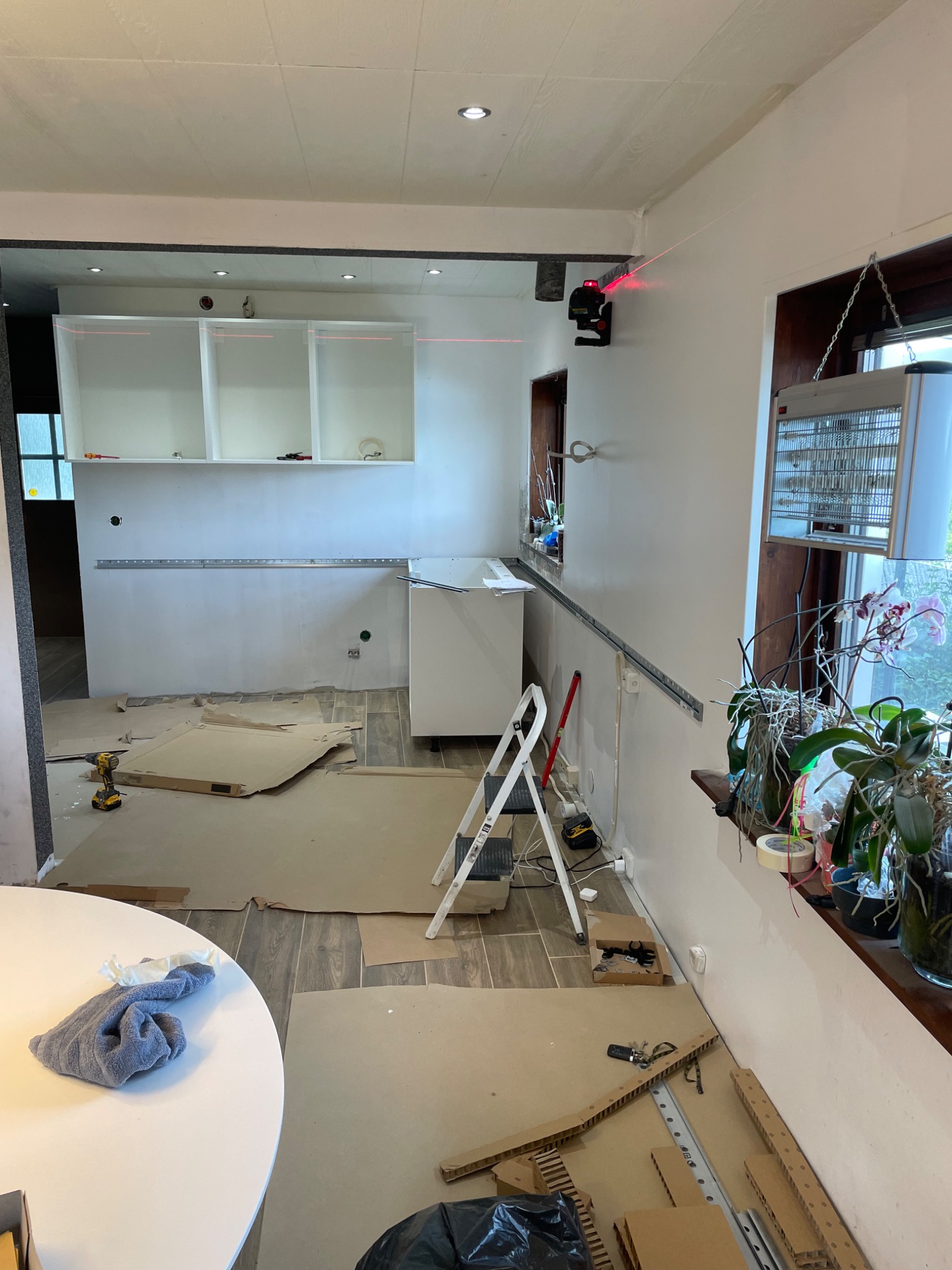 Wnętrze w trakcie montażu mebli kuchennych: widoczne białe szafki bez frontów, poziomica laserowa wyznaczająca linię, rozłożone kartony zabezpieczające podłogę, drabina i narzędzia.
