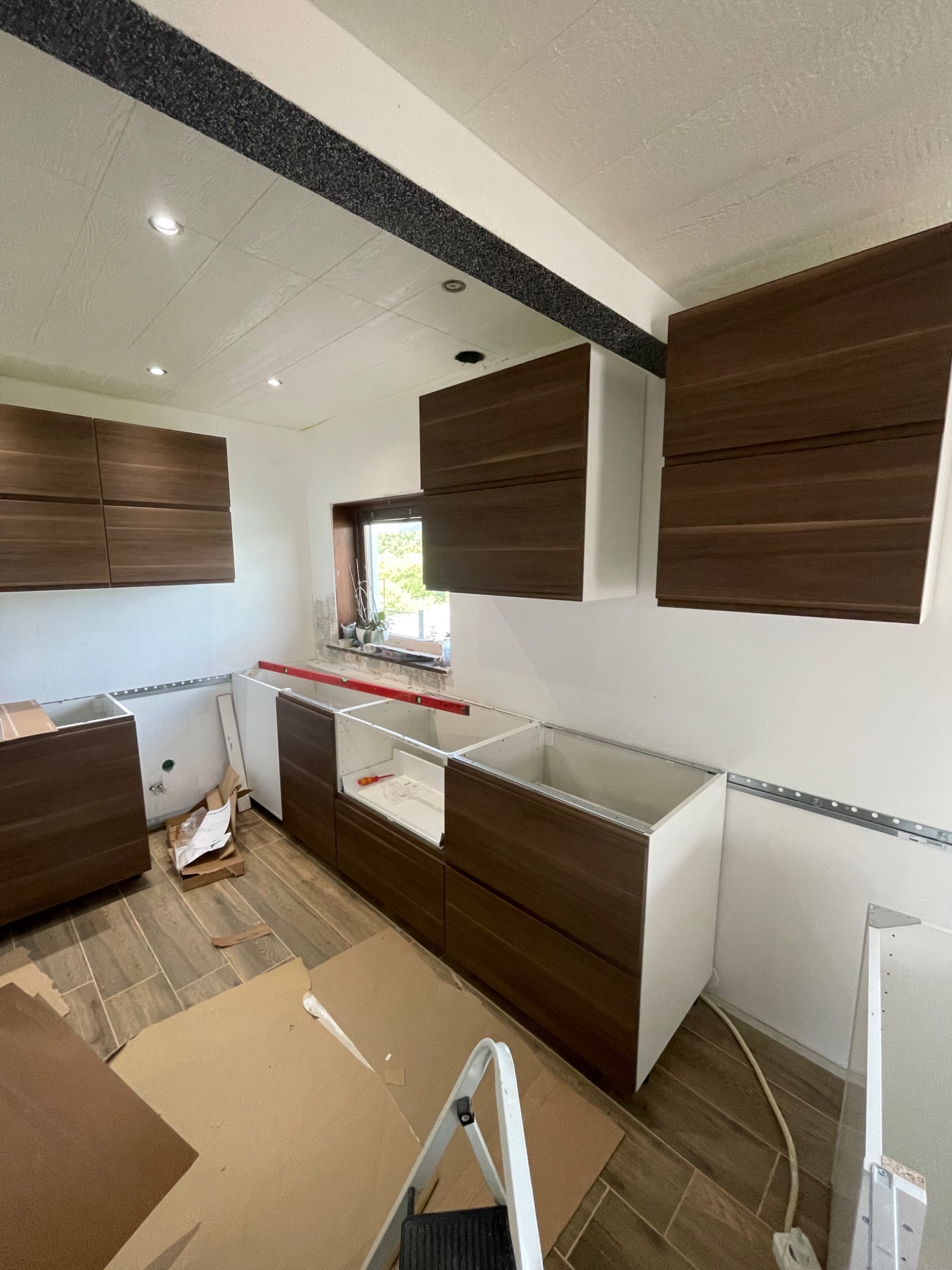 Montaż brązowo-białych szafek kuchennych z widoczną poziomicą i elementami montażowymi, na podłodze rozłożone kartony zabezpieczające.