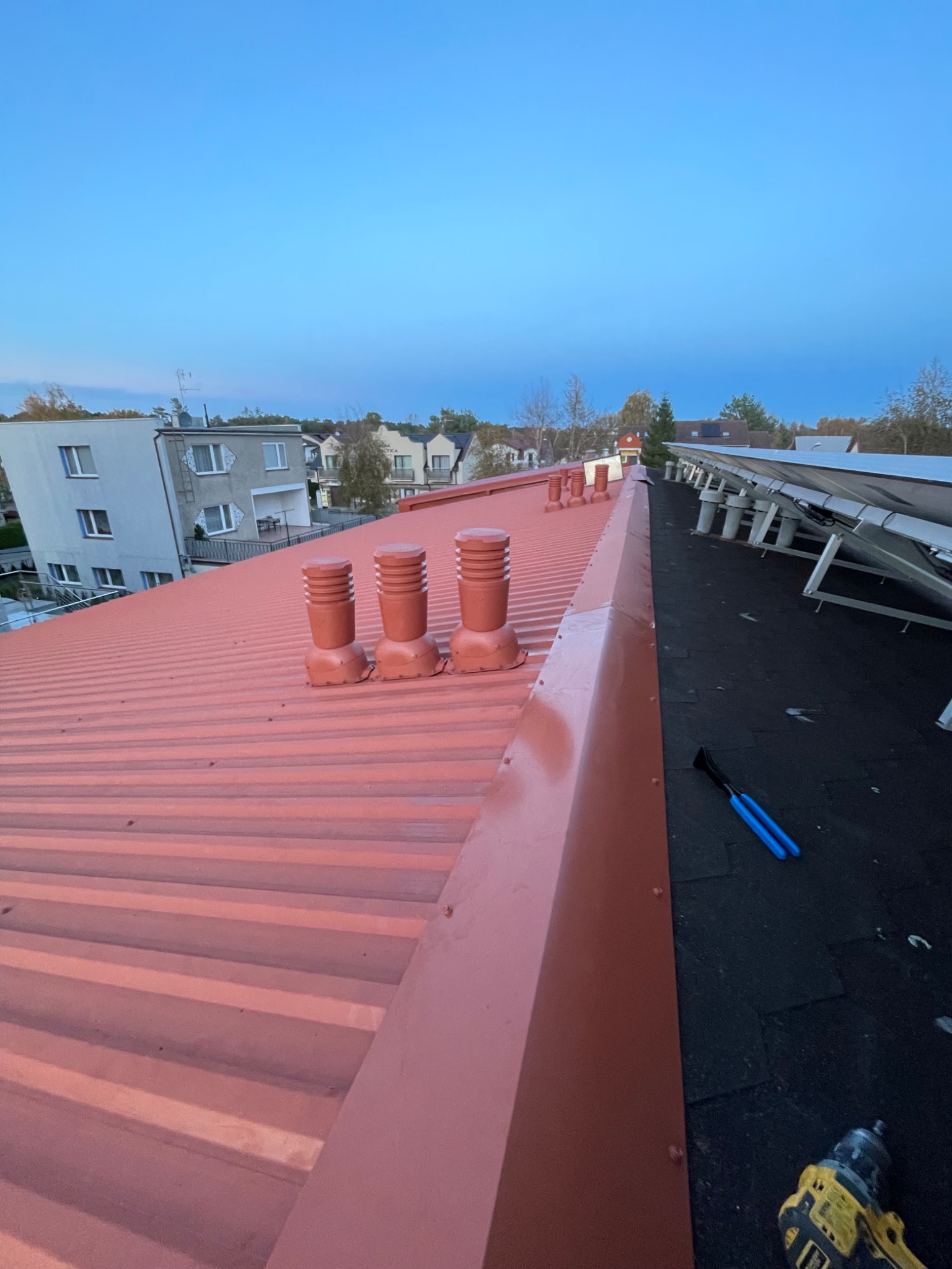 Czerwony blaszany dach z trzema kominami wentylacyjnymi i panelem słonecznym w tle, widoczne narzędzia dekarza: wkrętarka i szczypce.