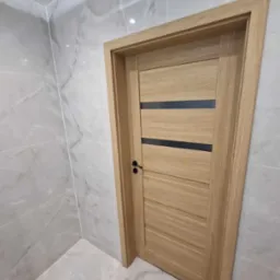 Jasne, drewniane drzwi wewnętrzne z czarnymi, poziomymi wstawkami, zamontowane w łazience wykończonej marmurowymi płytkami.