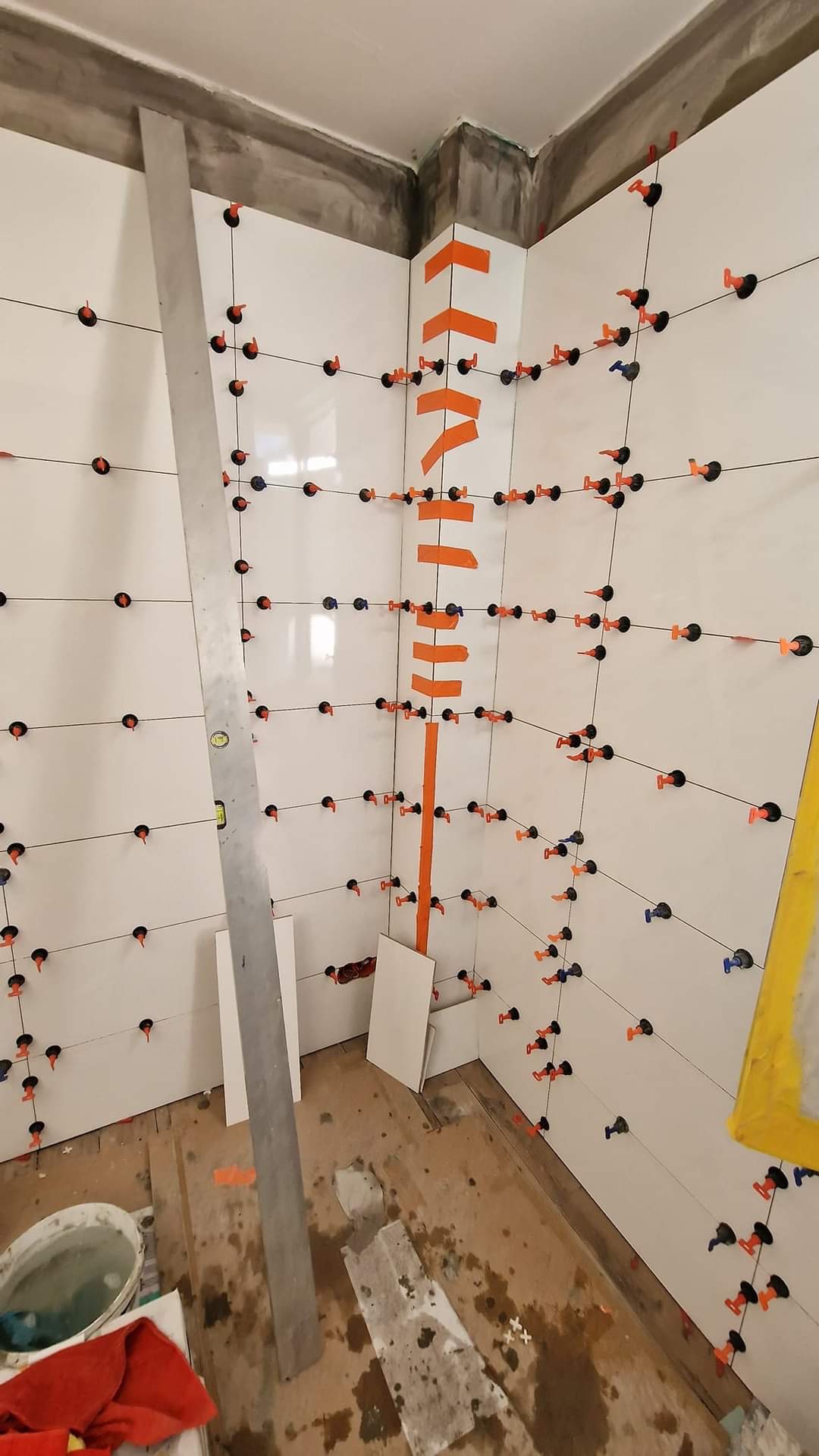 Układanie białych płytek w łazience z użyciem poziomicy, krzyżyków dystansowych i taśmy malarskiej wyznaczającej pion, widoczne narzędzia i wiadro.