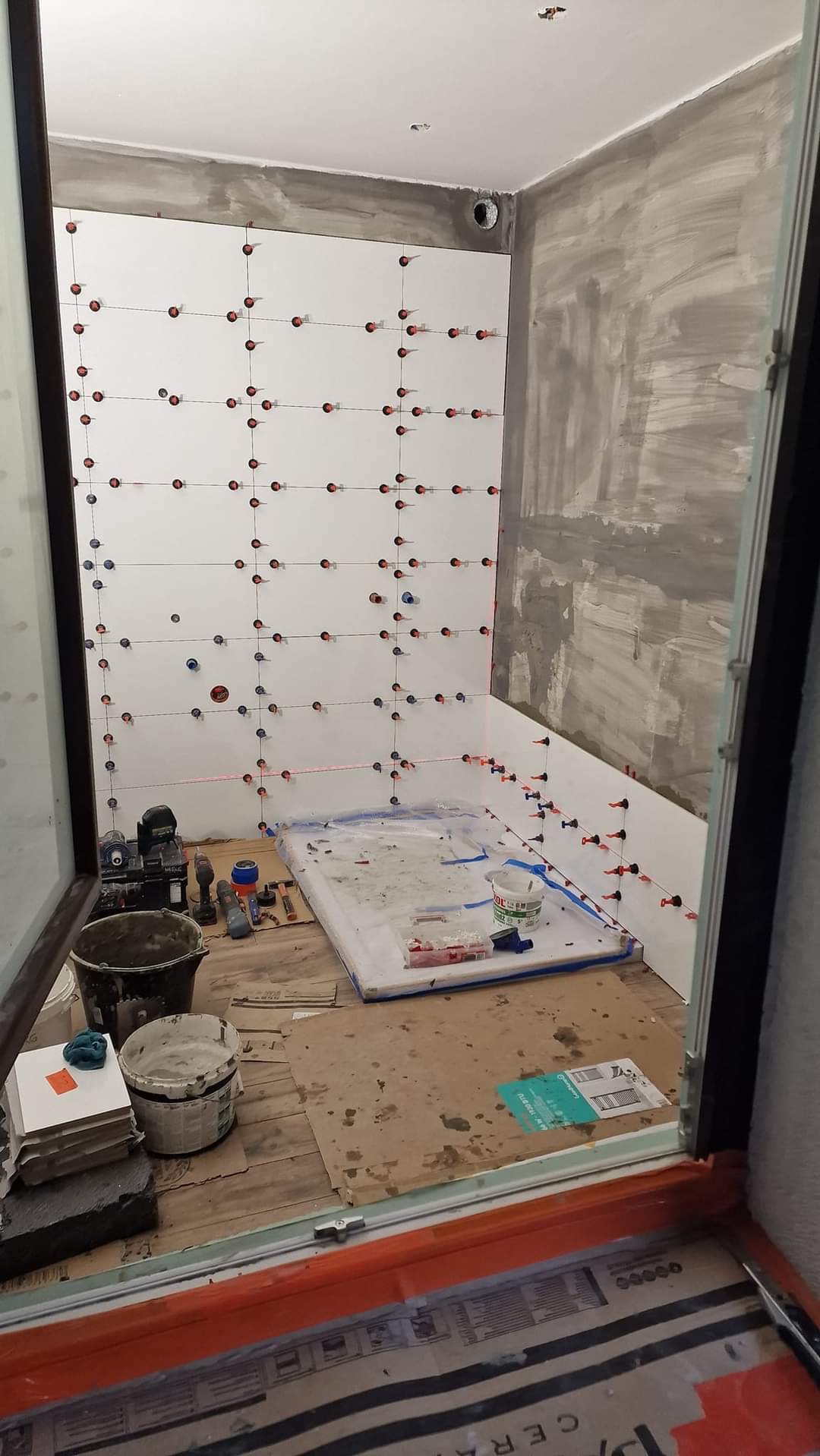 Łazienka w trakcie układania białych płytek na ścianach, z użyciem poziomicy laserowej i krzyżyków dystansowych, widoczne narzędzia i materiały budowlane.