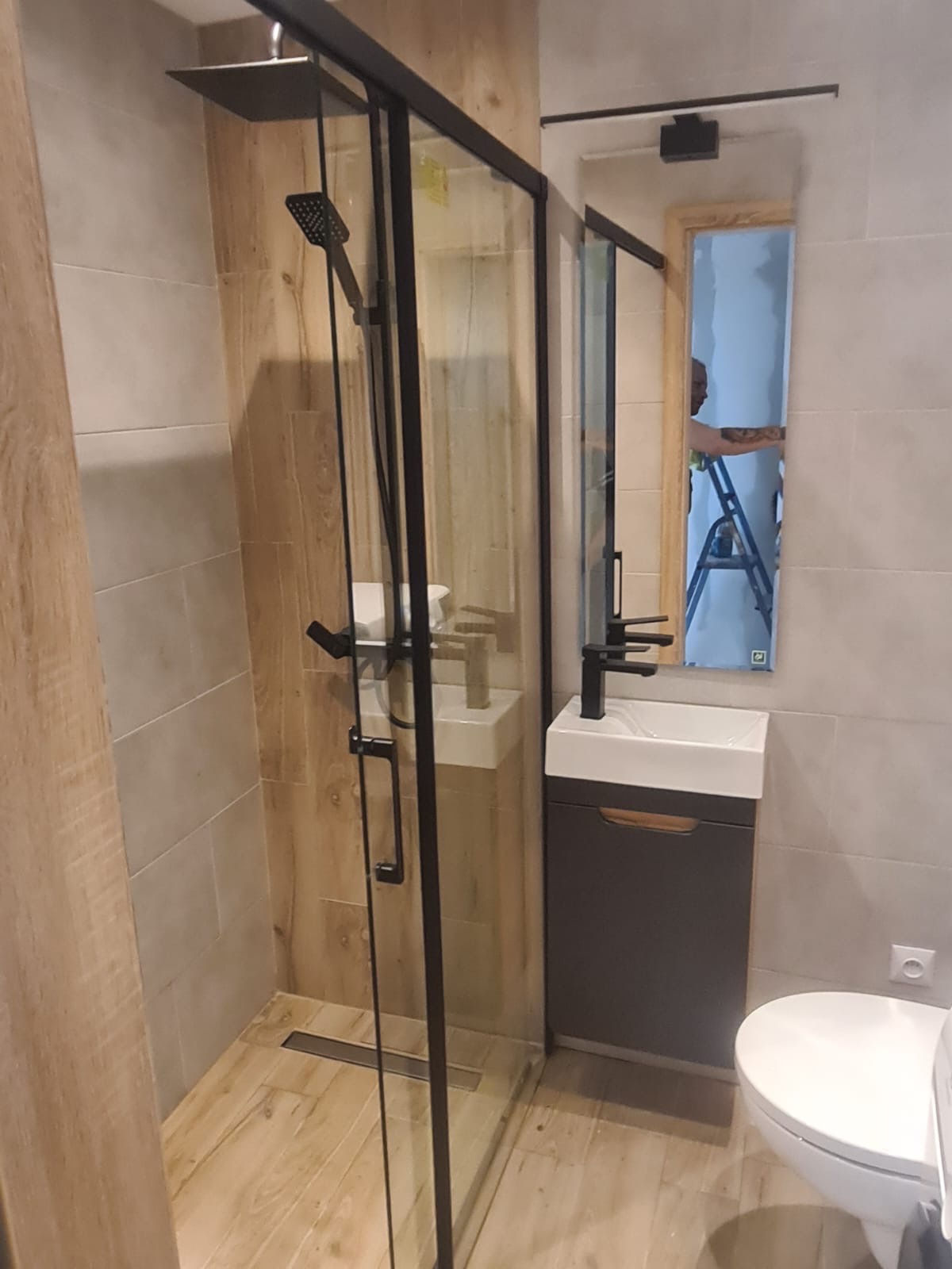 Nowoczesna łazienka z kabiną prysznicową typu walk-in z czarnymi profilami, umywalką nablatową na szafce w kolorze grafitowym i lustrem, w którym odbija się osoba na drabinie.