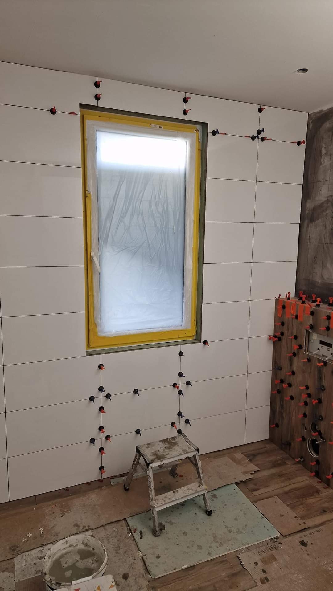 Układanie białych, poziomych płytek w łazience, z użyciem klipsów poziomujących, okno zabezpieczone folią, drabinka i wiadro z zaprawą na podłodze.