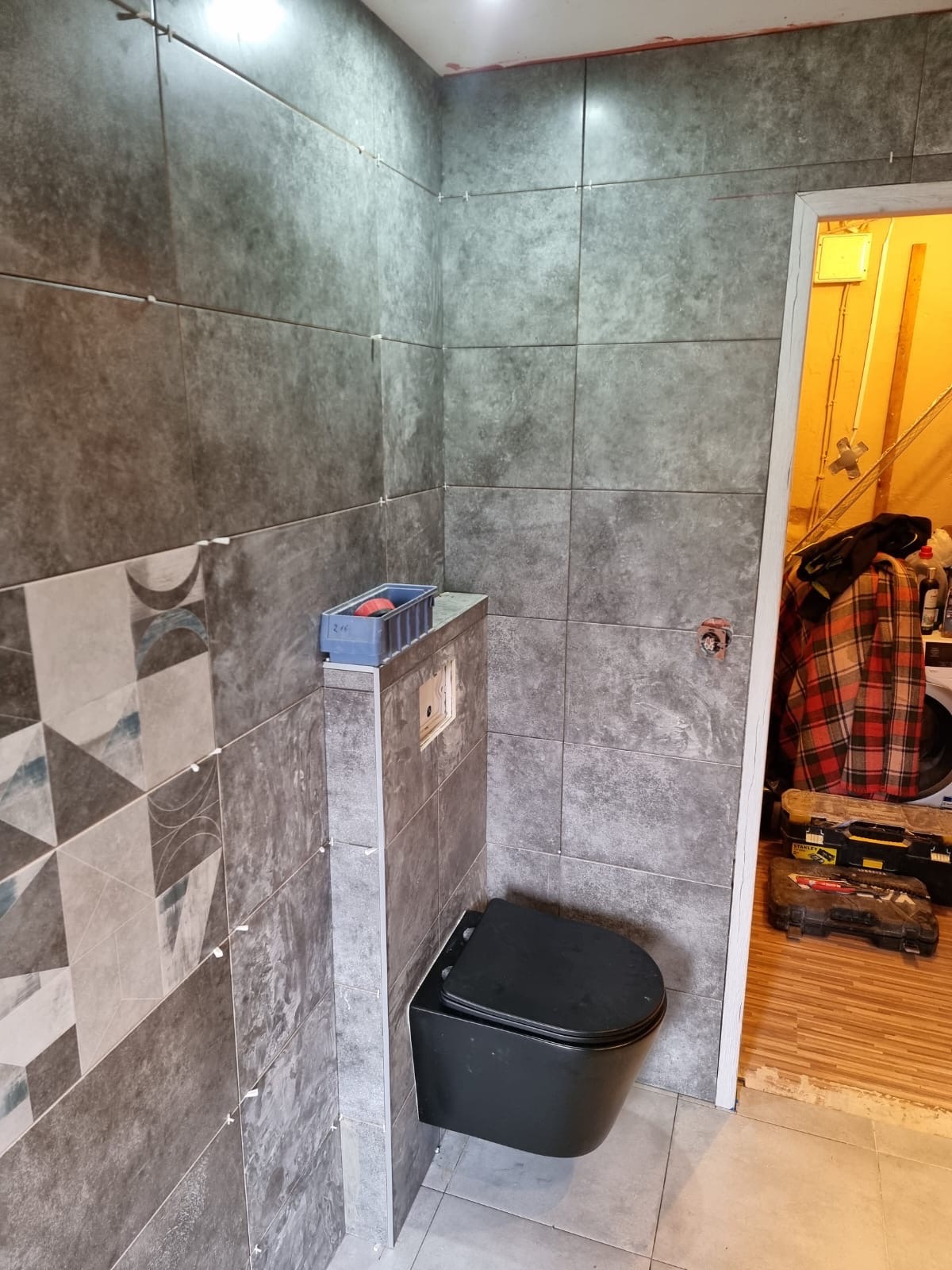 Wnętrze łazienki w trakcie remontu: szare płytki imitujące kamień na ścianach, czarna podwieszana toaleta, widoczne krzyżyki dystansowe, pojemnik z narzędziami na zabudowie stelaża, bałagan...