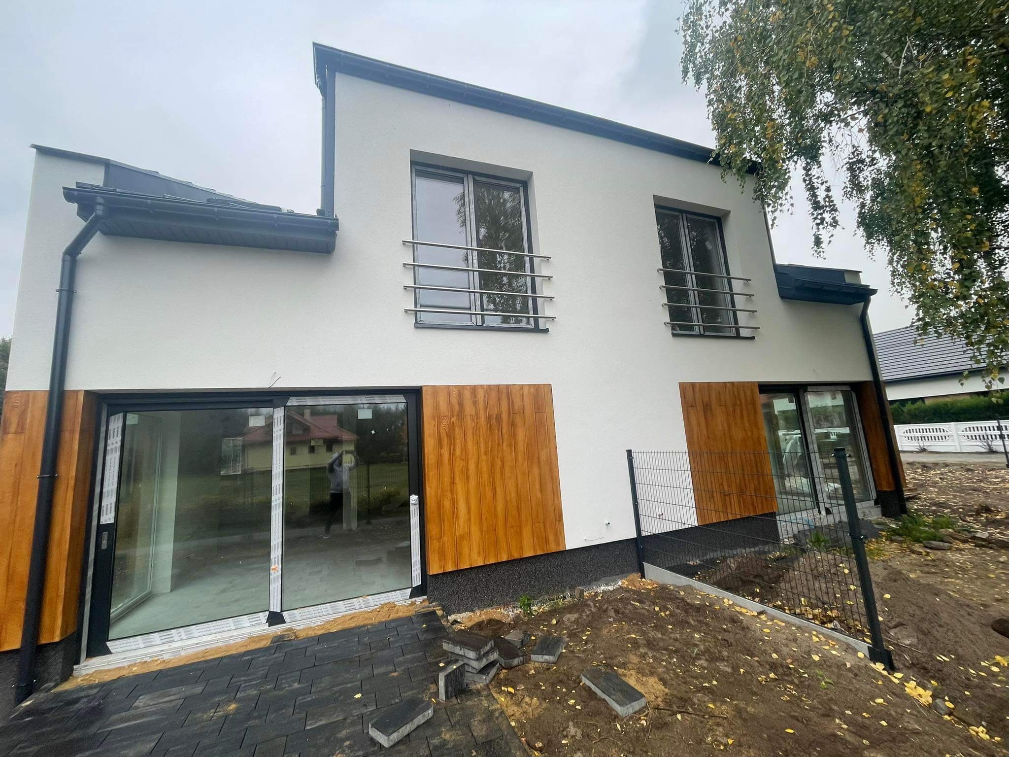 Nowoczesny dom z białymi ścianami, drewnianymi akcentami i metalowymi balustradami okiennymi, widoczne rozpoczęte prace brukarskie i montaż ogrodzenia panelowego.