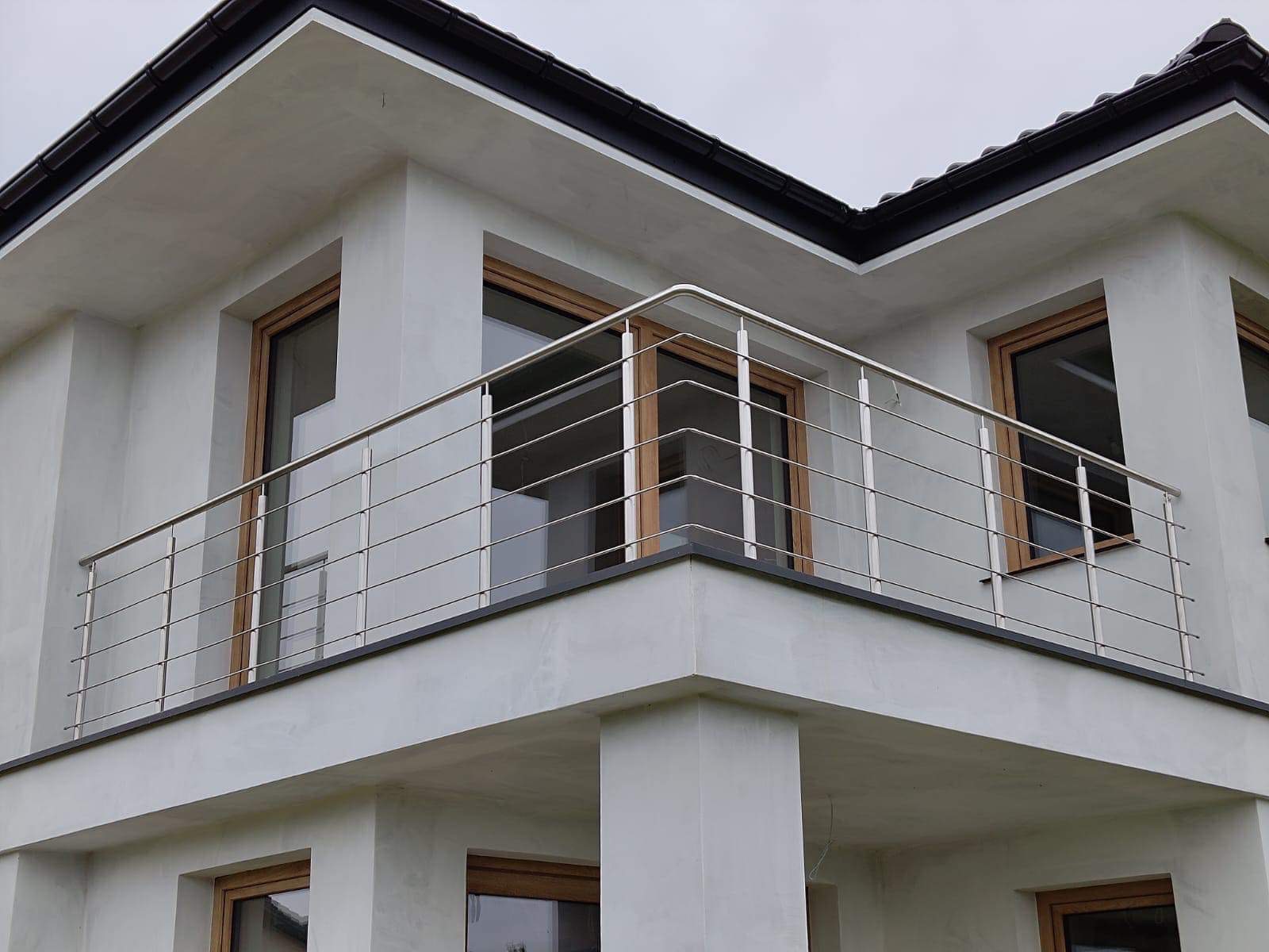 Nowoczesny balkon z prostą, poziomą balustradą ze stali nierdzewnej, widok z dołu na dwupiętrowy dom z oknami w drewnianych ramach.
