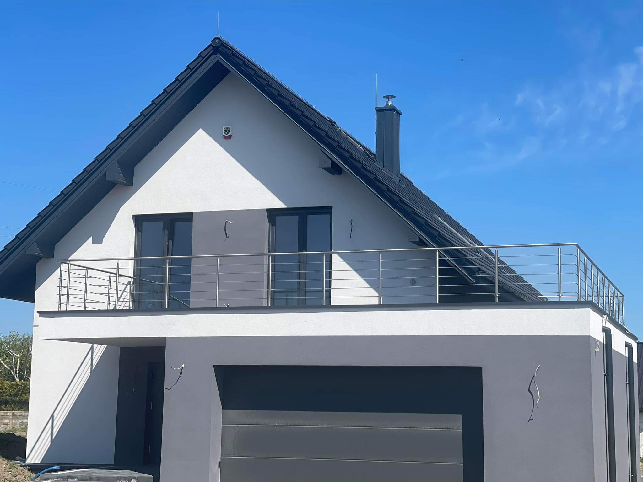 Nowoczesny dom jednorodzinny z balkonem zabezpieczonym minimalistyczną, metalową barierką, widok z zewnątrz w słoneczny dzień.