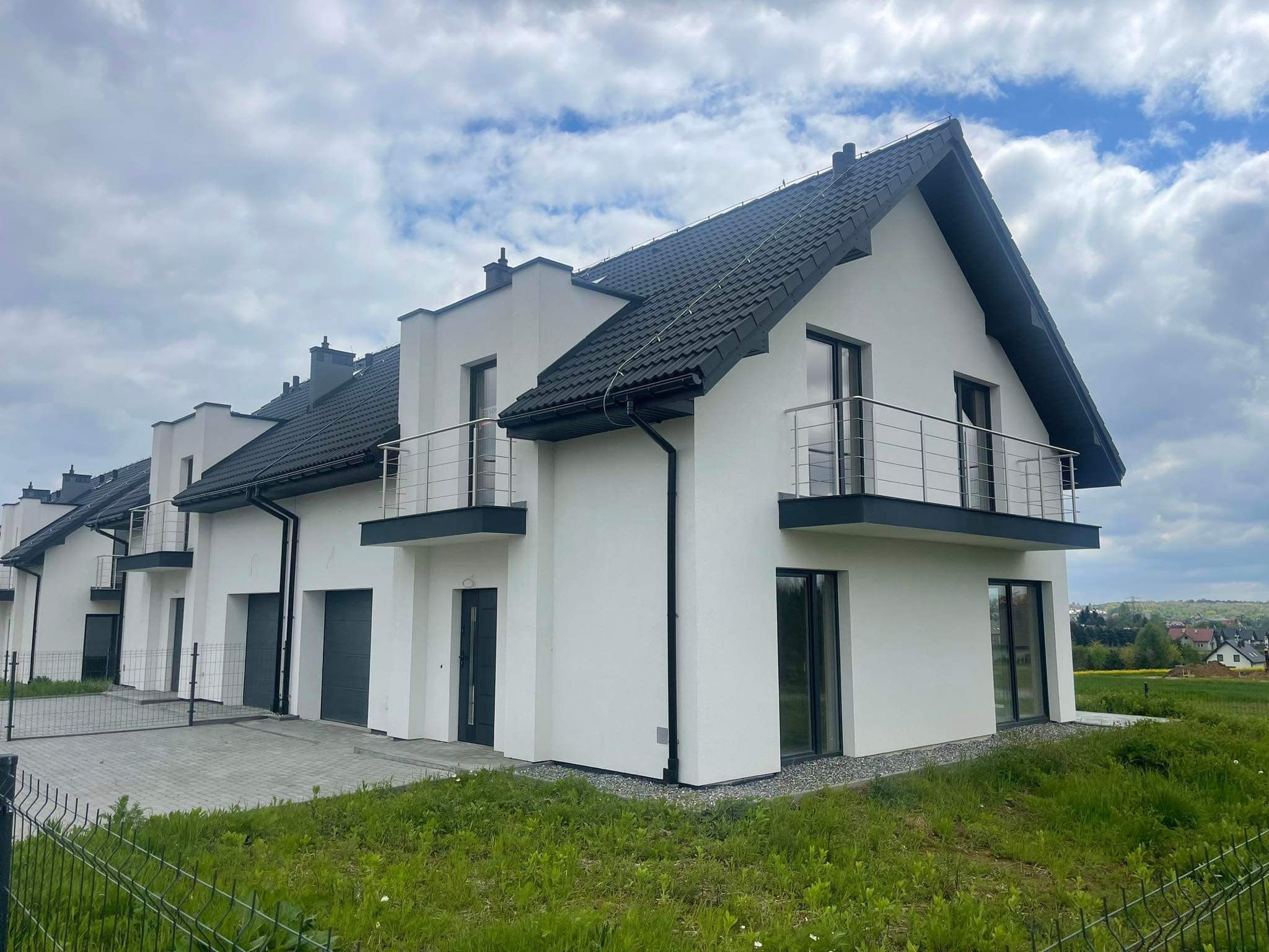 Szereg domów jednorodzinnych z balkonami z metalowymi barierkami, wykończonych w nowoczesnym stylu, z widocznymi elementami drenażu i terenem zielonym przed budynkami.
