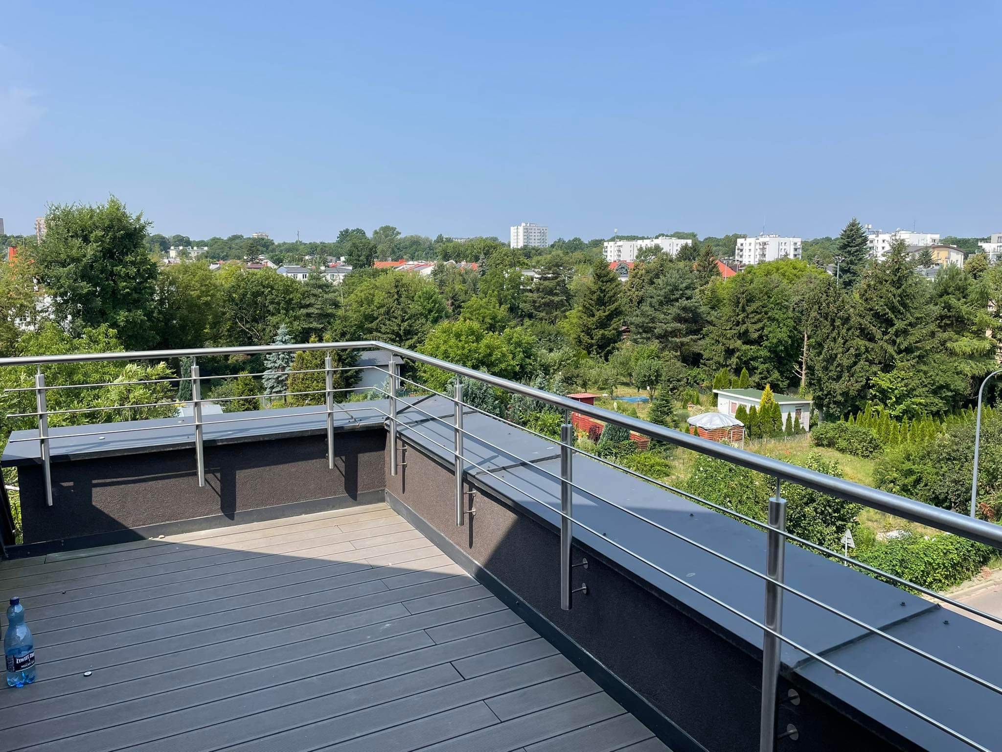 Nowoczesny balkon z szarą podłogą z desek kompozytowych i stalową balustradą z widokiem na zielone drzewa i zabudowania w tle.