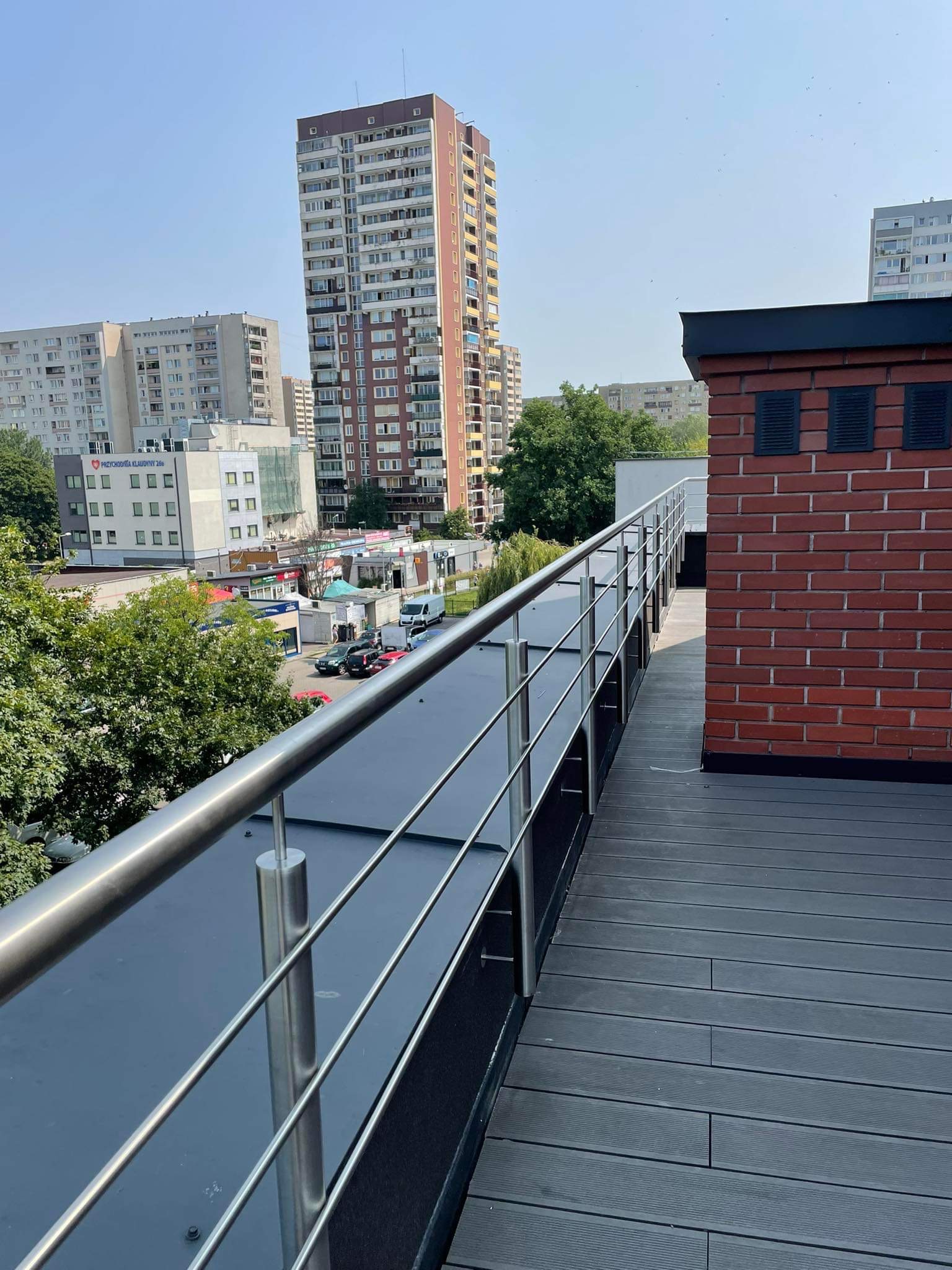 Balkon z szarą podłogą kompozytową i prostą, metalową balustradą na tle miejskiego krajobrazu z wysokimi budynkami.