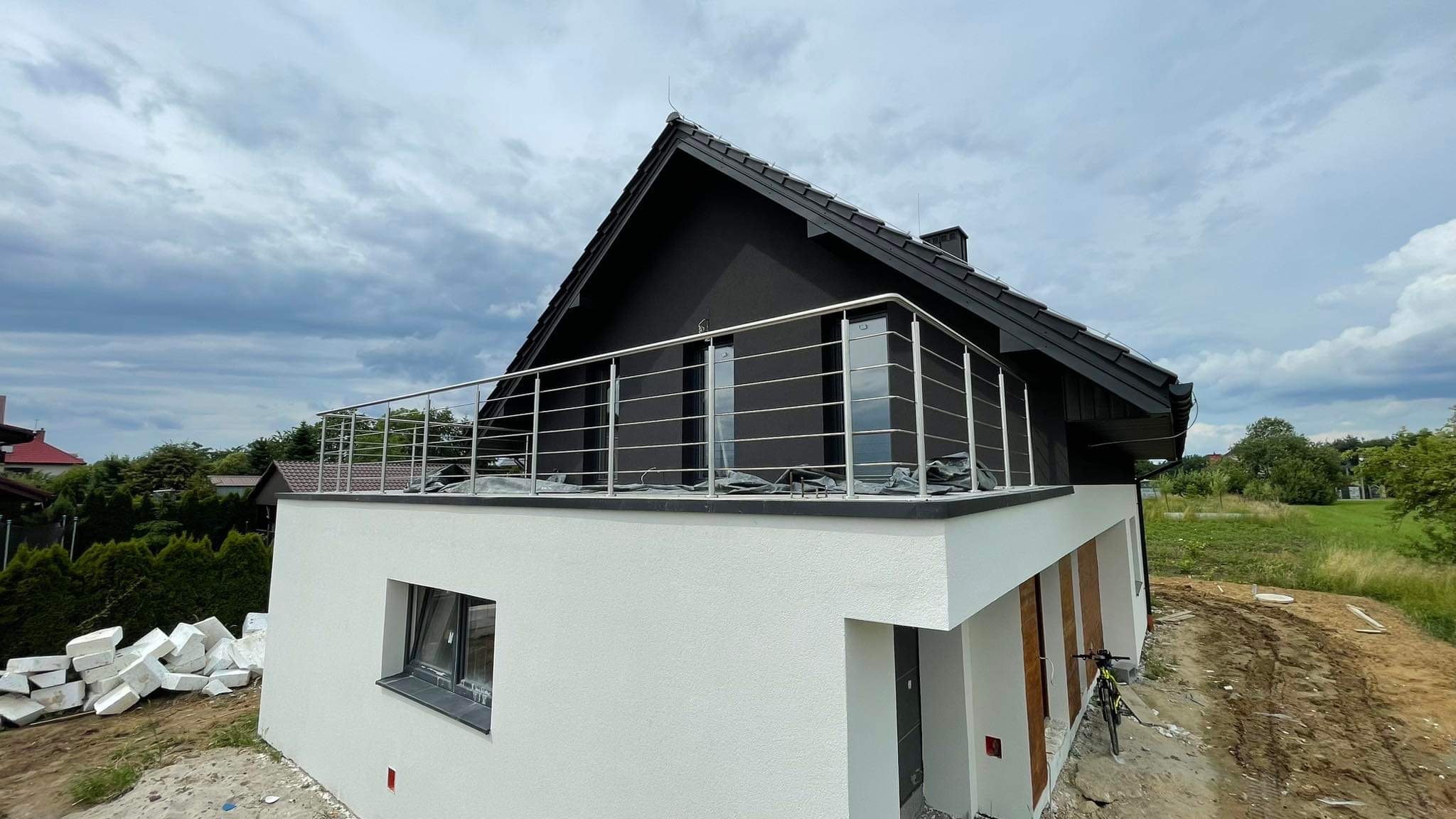 Nowoczesny dom z czarną elewacją i balkonem zabezpieczonym metalową, minimalistyczną barierką ze stali nierdzewnej, widok z dołu na tle pochmurnego nieba.