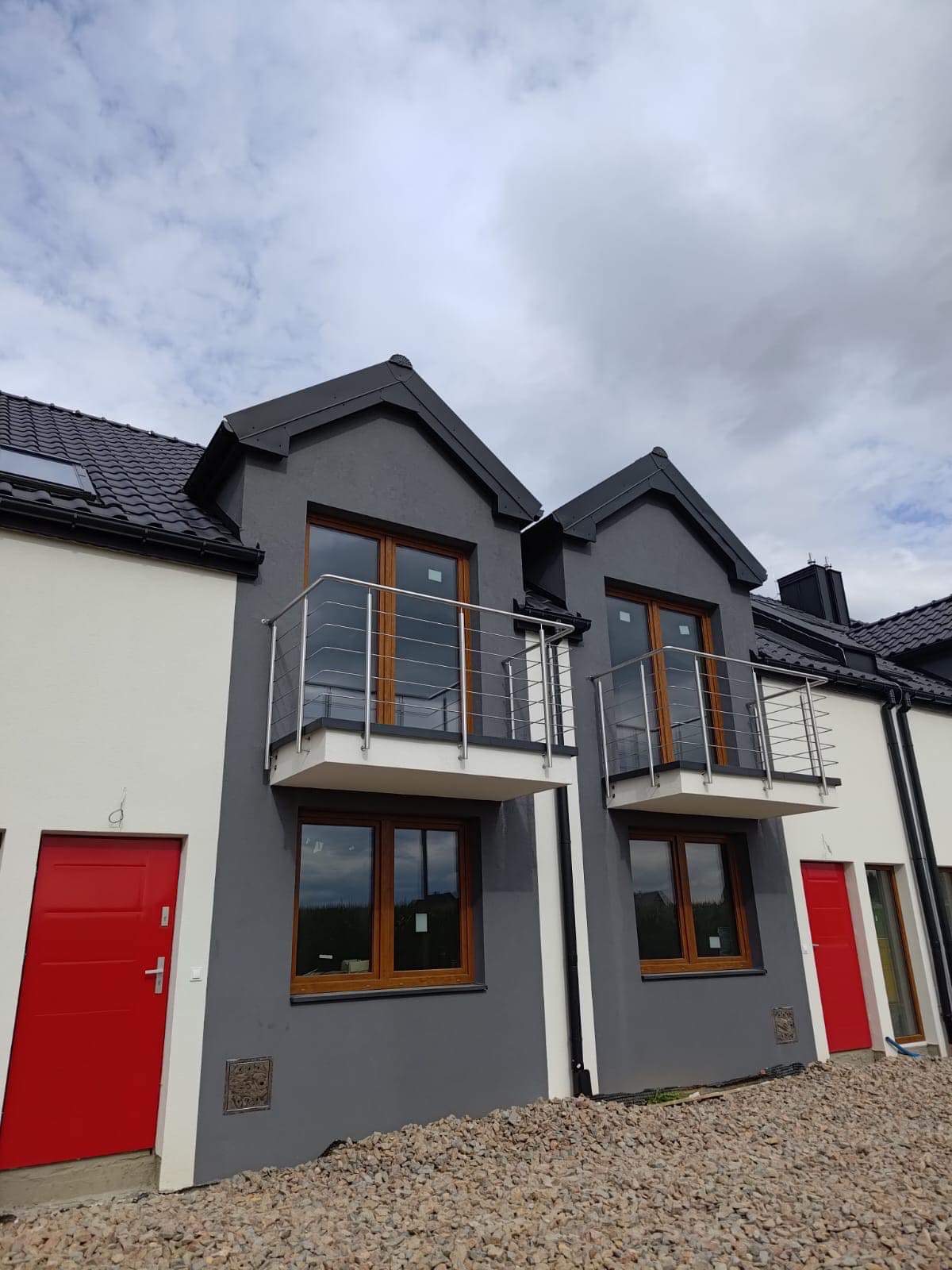 Dwa balkony z balustradami nierdzewnymi w nowym domu z szarą elewacją i czerwonymi drzwiami wejściowymi, widok z dołu na tle pochmurnego nieba.