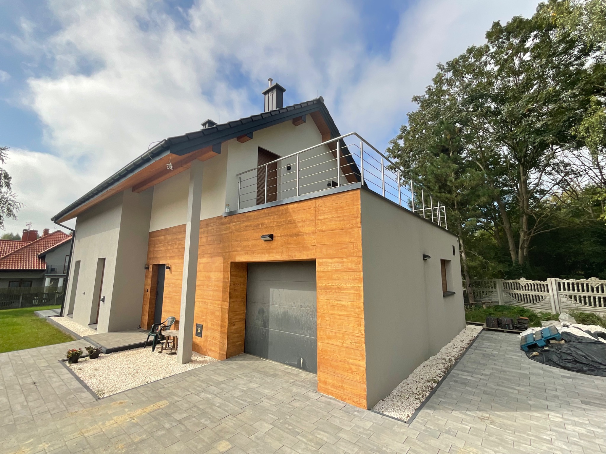 Nowoczesny dom jednorodzinny z balkonem zabezpieczonym stalową balustradą poziomą, widok z poziomu brukowanego podjazdu, błękitne niebo z chmurami w tle.