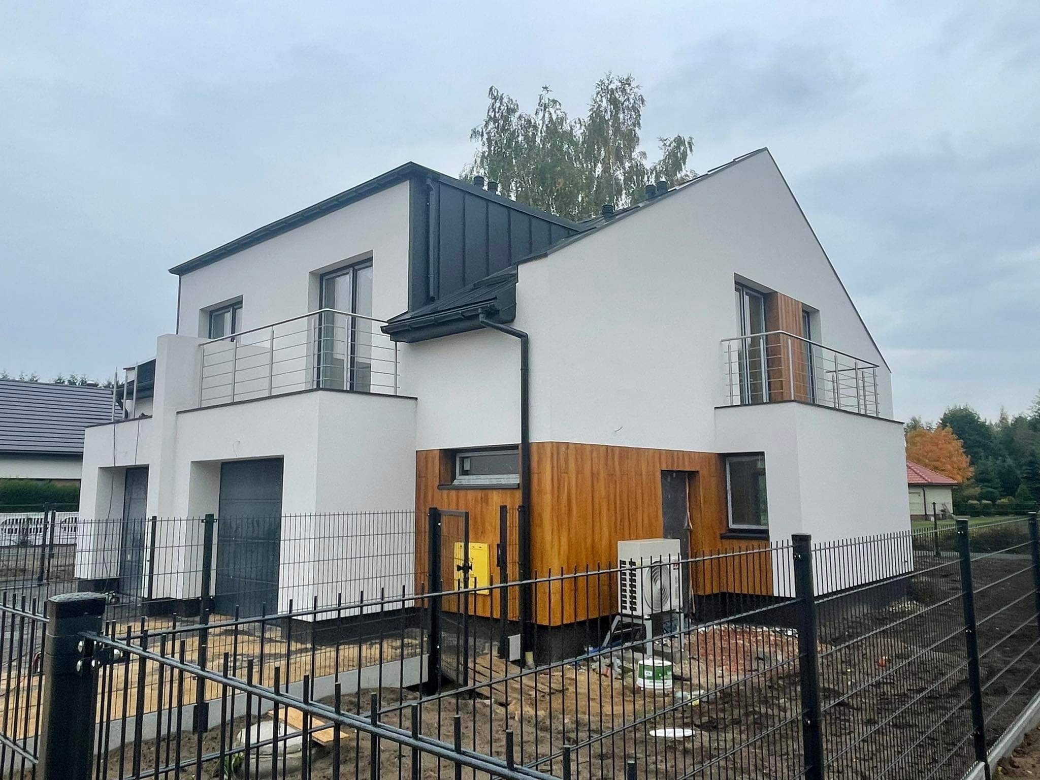 Nowoczesny dom jednorodzinny z balkonami z prostymi, stalowymi barierkami, wykończony drewnianą elewacją i otoczony czarnym, metalowym ogrodzeniem panelowym na etapie montażu.