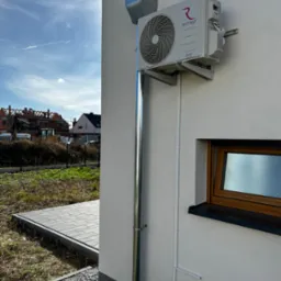 Zewnętrzna jednostka klimatyzacji Rotenso Roni Series zamontowana na ścianie domu, z widocznymi rurami odprowadzającymi skropliny i przewodami elektrycznymi w białych listwach, na tle jasnej...