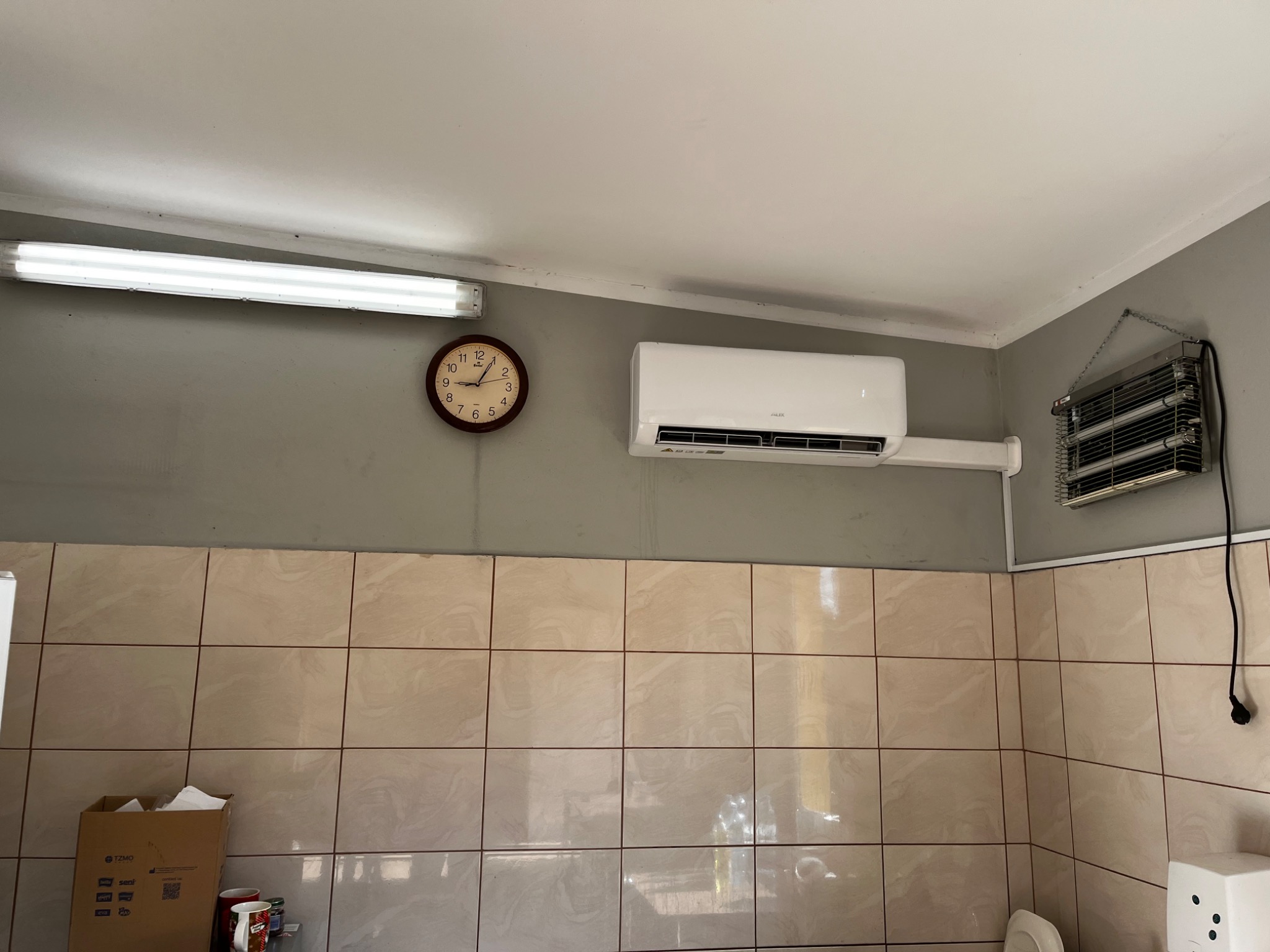 Biały klimatyzator ścienny zamontowany w pomieszczeniu z beżowymi płytkami, widoczny zegar ścienny i lampa fluorescencyjna.