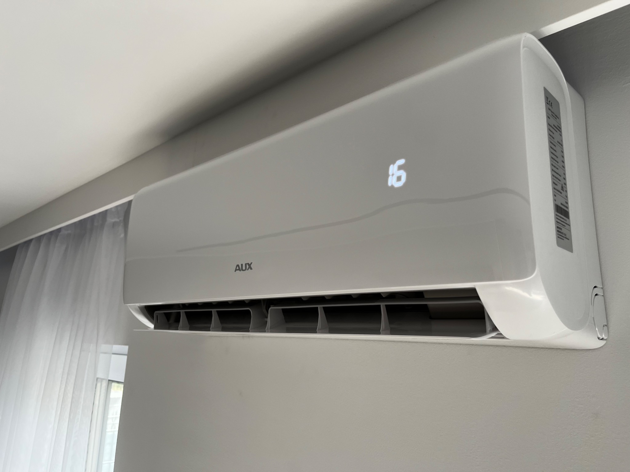 Biała klimatyzacja marki AUX zamontowana na ścianie, wyświetlająca temperaturę 16 stopni, widoczne zasłony w tle.