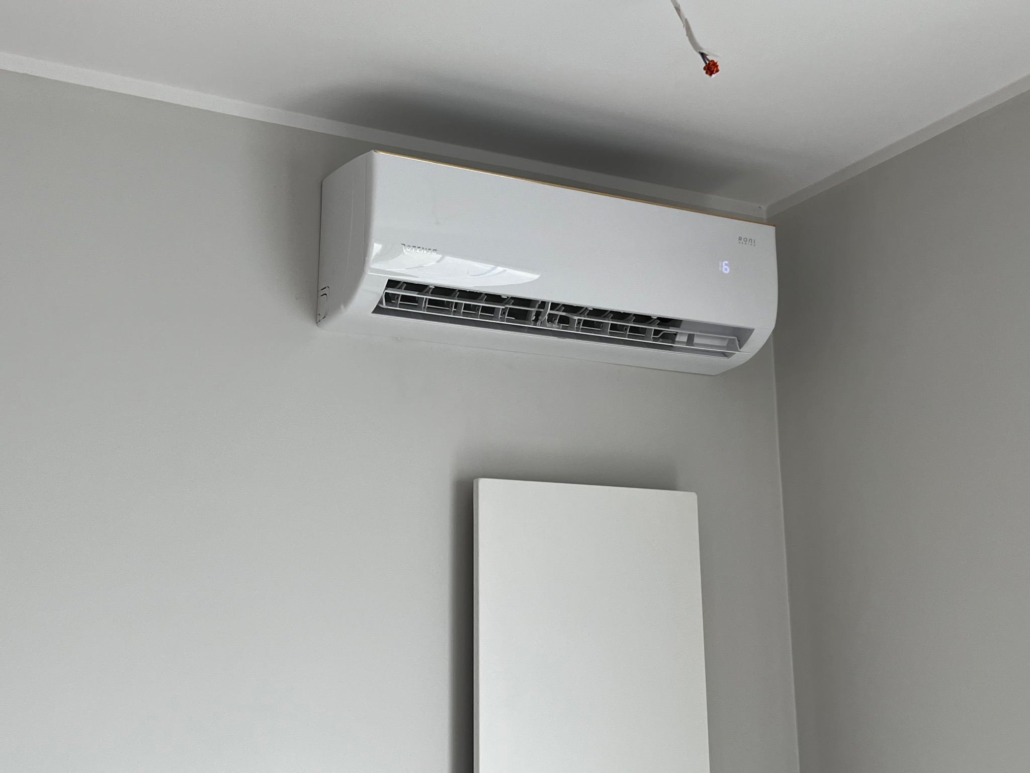 Nowoczesny biały klimatyzator z eleganckim złotym paskiem u góry, zamontowany na szarej ścianie w minimalistycznym wnętrzu, widoczny przewód elektryczny w rogu sufitu.