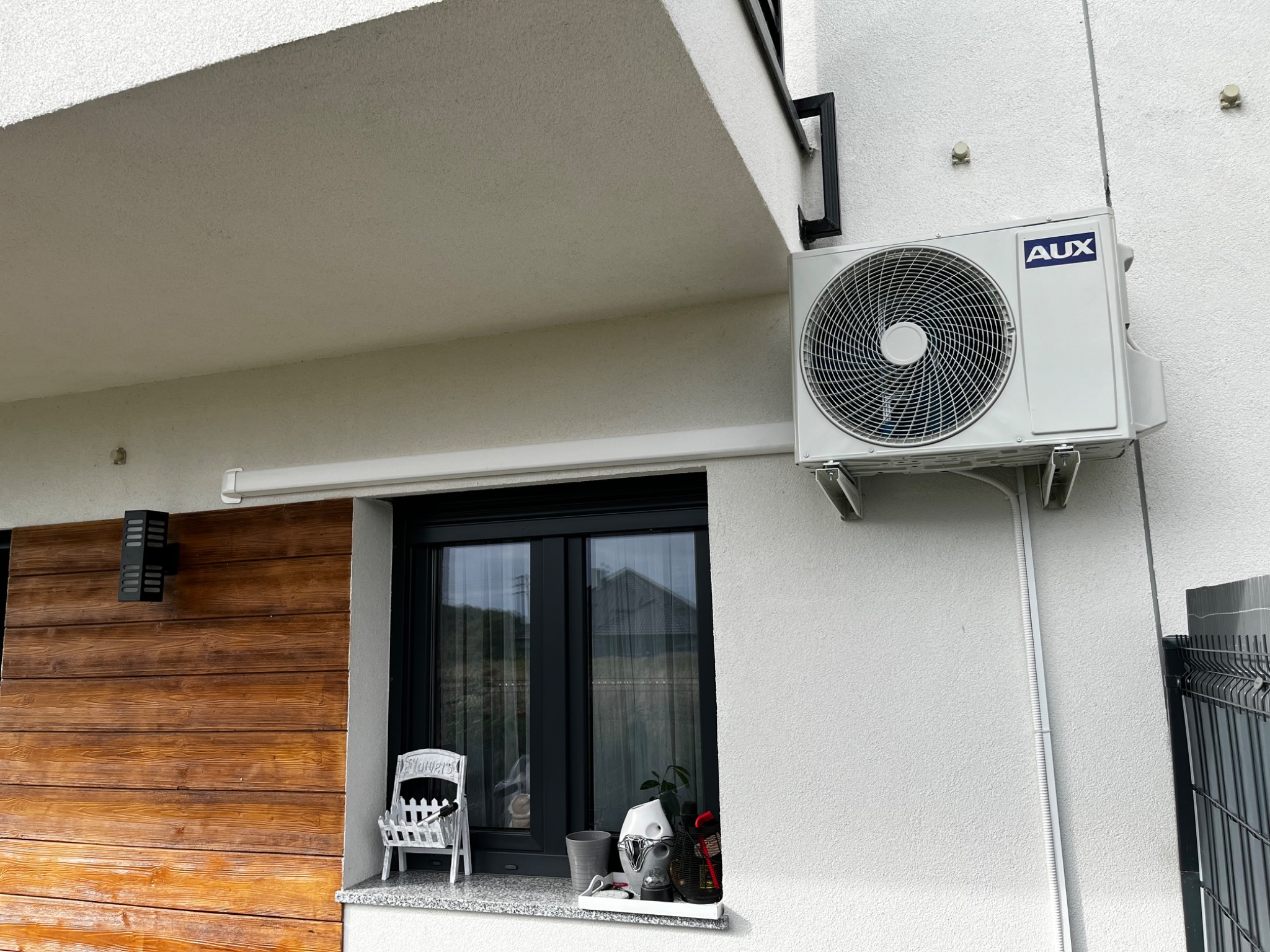 Biały klimatyzator marki AUX zamontowany na elewacji domu z drewnianą okładziną i czarną ramą okienną, widoczne prowadnice kabli i dekoracje okienne