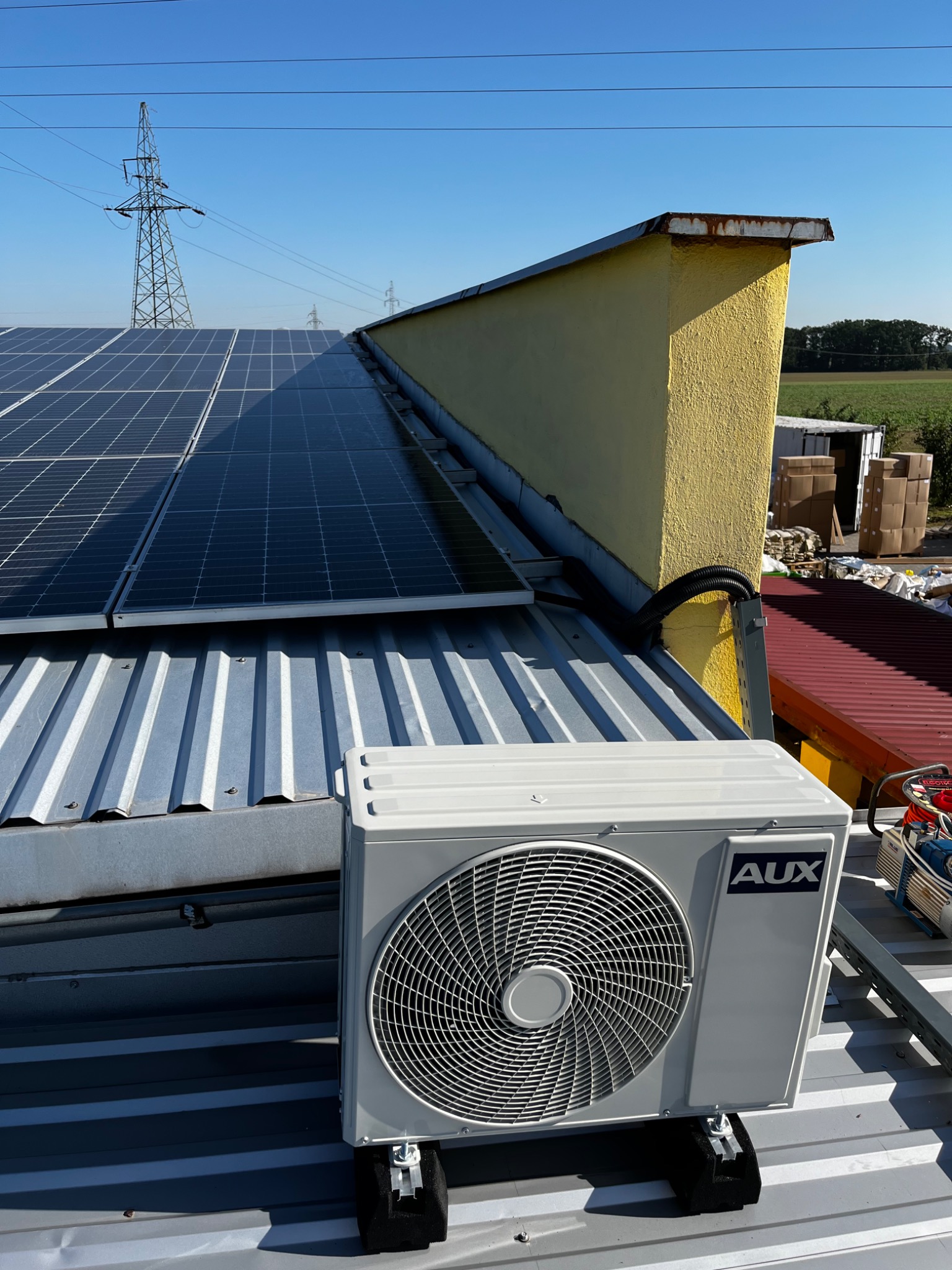 Zewnętrzna jednostka klimatyzacji AUX zamontowana na metalowym dachu budynku z panelami słonecznymi, widoczna żółta ściana i słup wysokiego napięcia w tle.