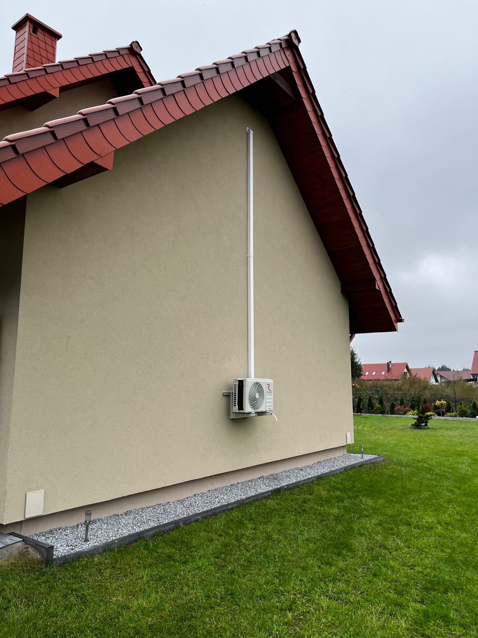 Zewnętrzna jednostka klimatyzacji zamontowana na elewacji domu jednorodzinnego z widocznymi rurami odprowadzającymi skropliny i osłoną instalacji, na tle zadbanego trawnika i szarego nieba.