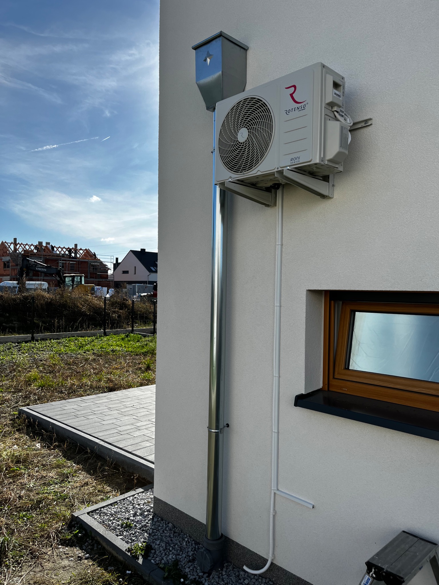 Zewnętrzna jednostka klimatyzacji Rotenso Roni Series zamontowana na ścianie domu, z widocznymi rurami odprowadzającymi skropliny i przewodami elektrycznymi w białych listwach, na tle jasnej...