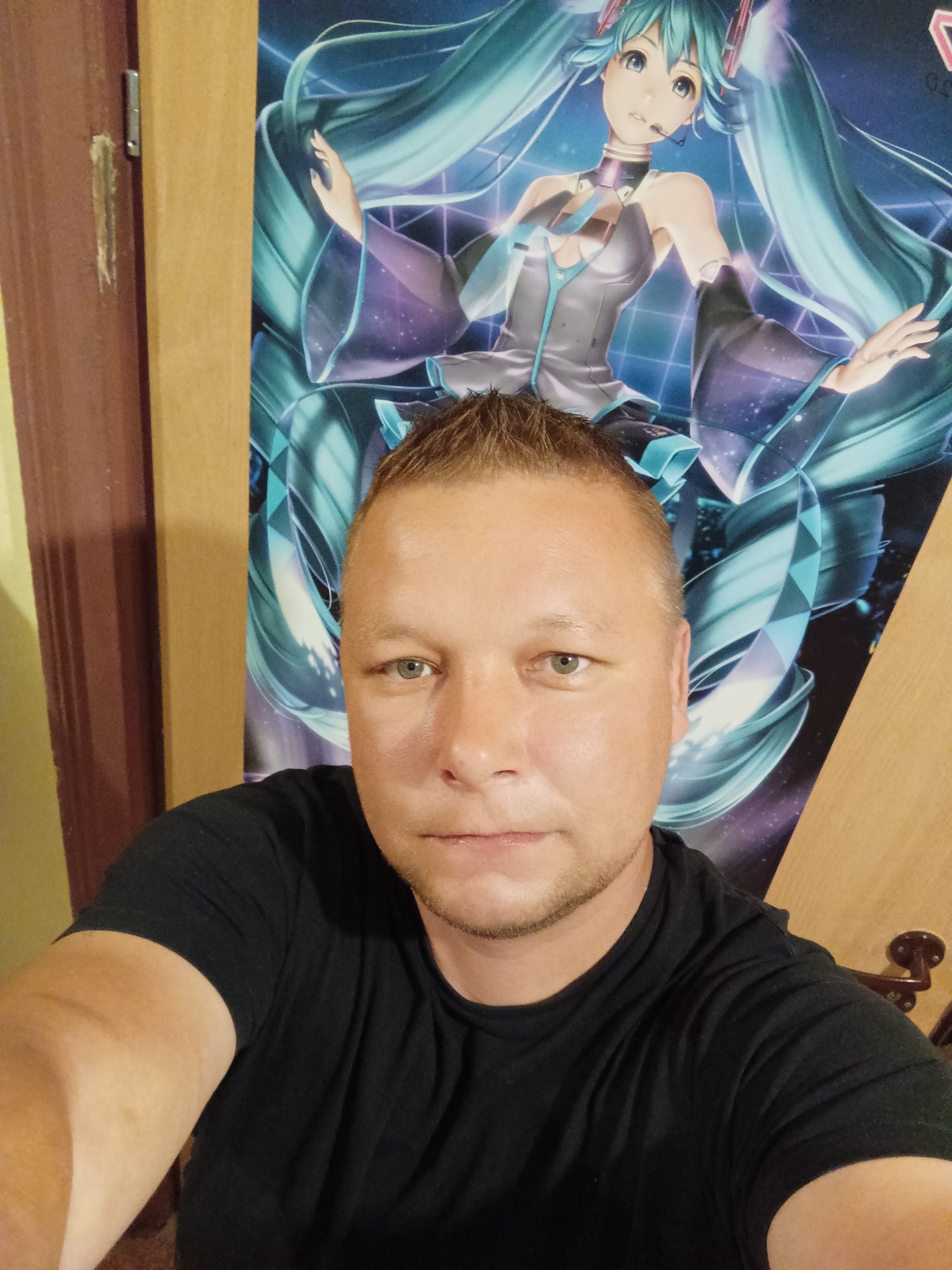 Mężczyzna w czarnej koszulce robiący selfie na tle plakatu z anime Hatsune Miku, widoczne ślady po usunięciu czegoś ze ściany.