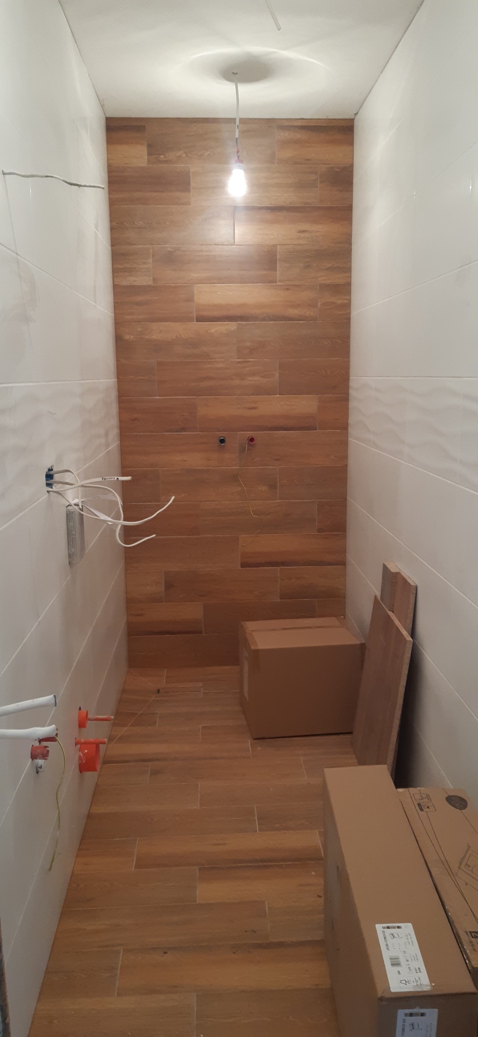 Wąska łazienka w trakcie prac wykończeniowych: białe płytki ścienne, drewnopodobne płytki na podłodze i ścianie, widoczne instalacje elektryczne i hydrauliczne, kartony z materiałami budowlanymi.