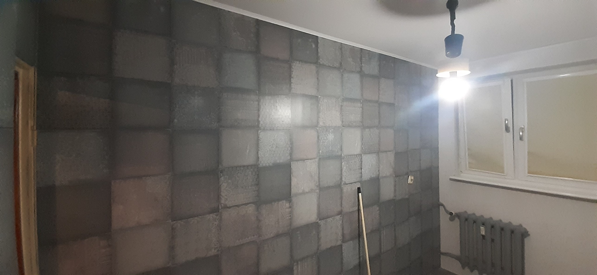 Ściana pokryta tapetą w kwadraty w odcieniach szarości i beżu, widoczny biały sufit z lampą wiszącą, okno z grzejnikiem i fragment drewnianej listwy przypodłogowej