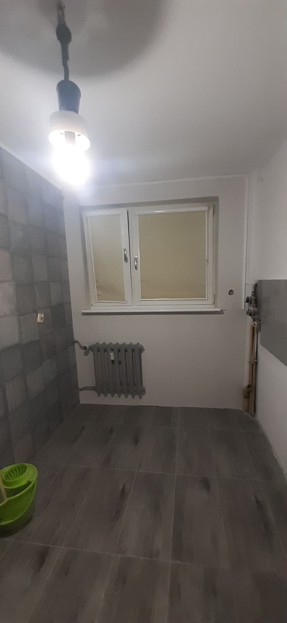 Wnętrze pokoju z szarą, wzorzystą ścianą, grzejnikiem pod oknem, podłogą wyłożoną płytkami imitującymi drewno i wiszącą żarówką.