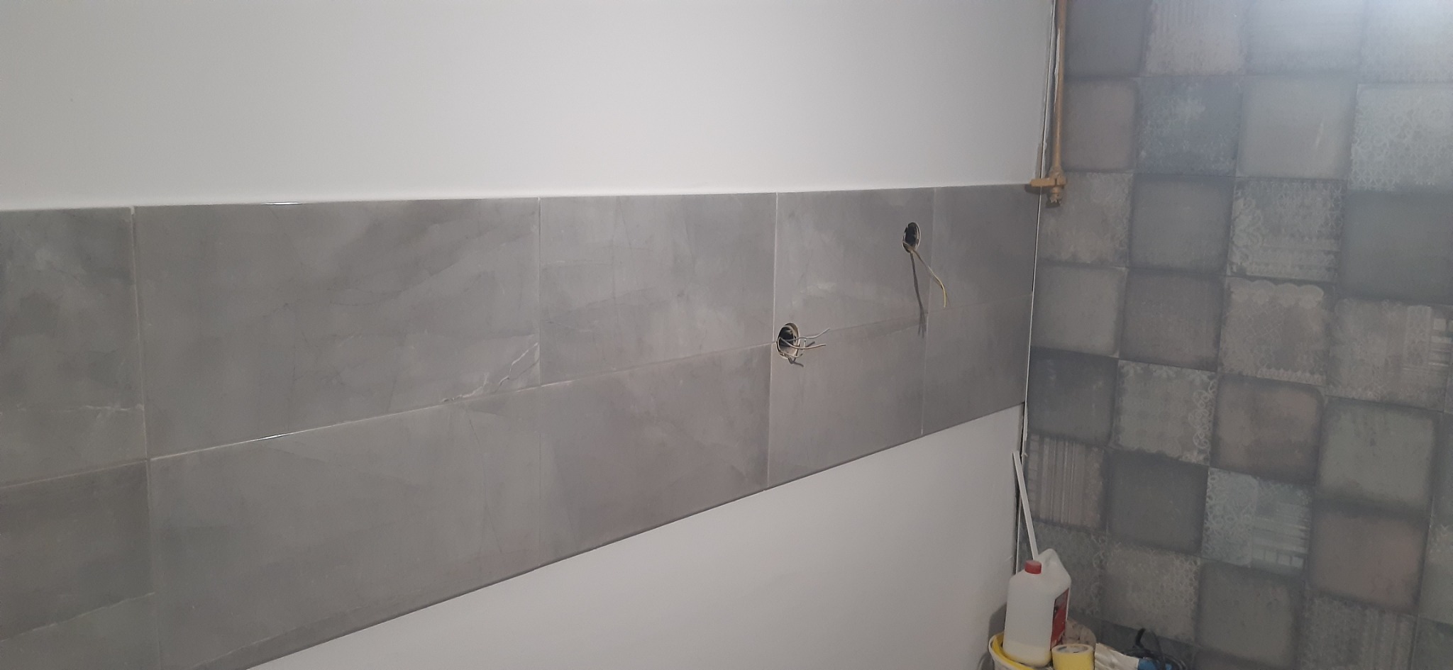 Ściana w trakcie remontu łazienki z ułożonymi szarymi płytkami imitującymi marmur, widoczne otwory na gniazdka elektryczne z wystającymi kablami oraz fragment ściany z dekoracyjnymi płytkami...