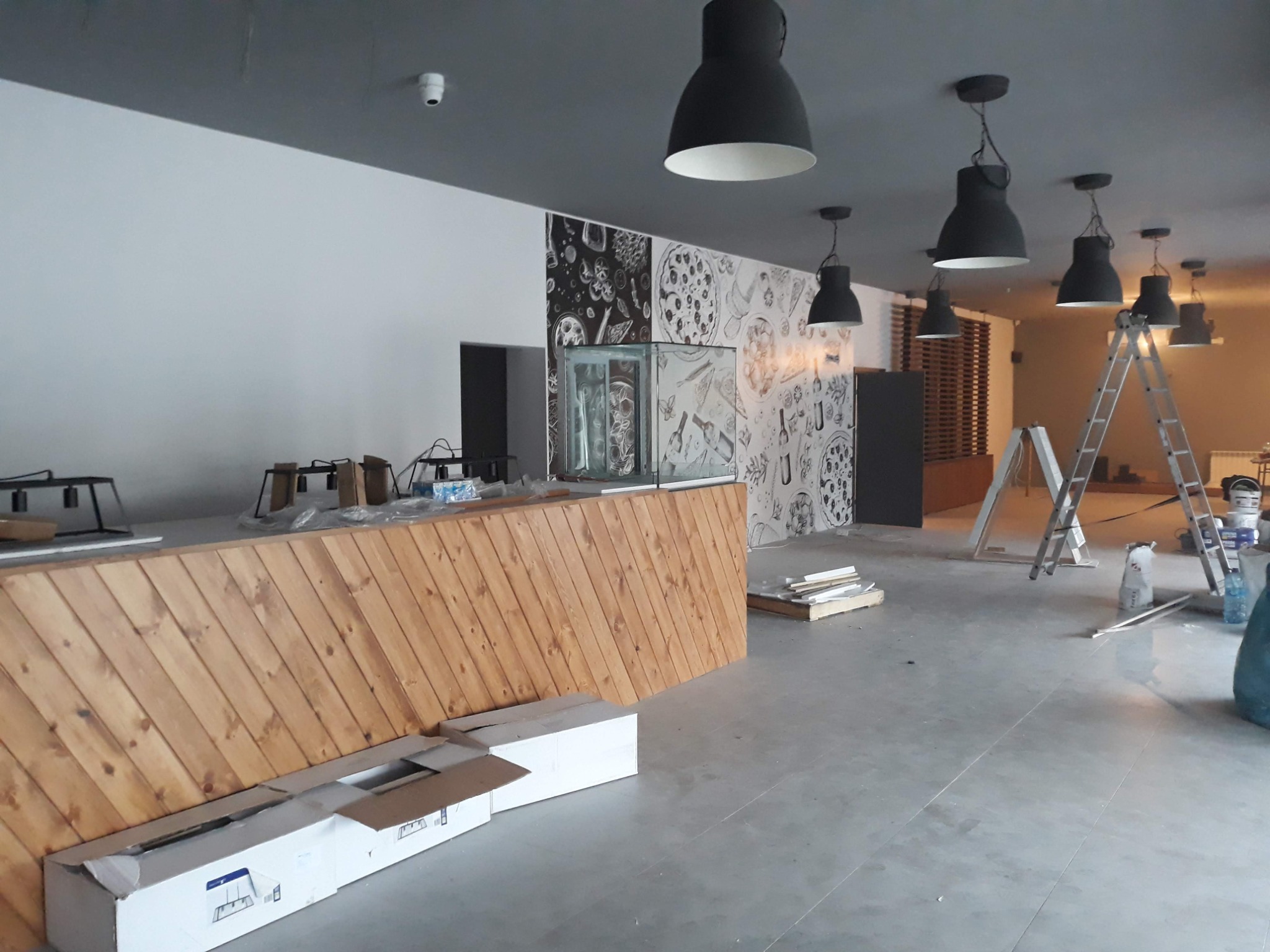 Wnętrze lokalu gastronomicznego w trakcie prac wykończeniowych: drewniana lada z ukośnych desek, ściana z motywami pizzy, czarne lampy przemysłowe, drabiny malarskie, kartony i materiały budowlane...