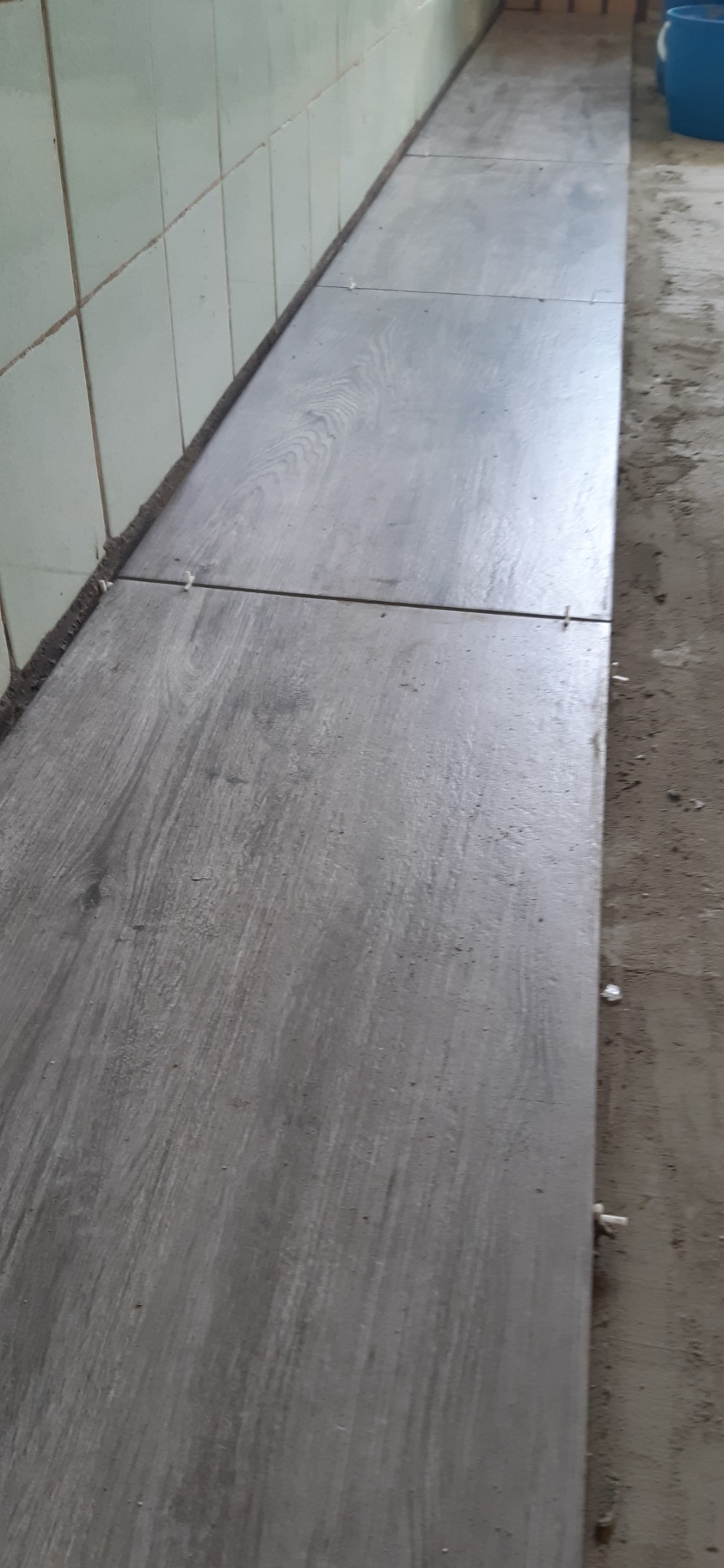 Układanie podłogi z szarych płytek imitujących drewno, widoczne plastikowe krzyżyki dystansowe między płytkami, przy ścianie z jasnych płytek.