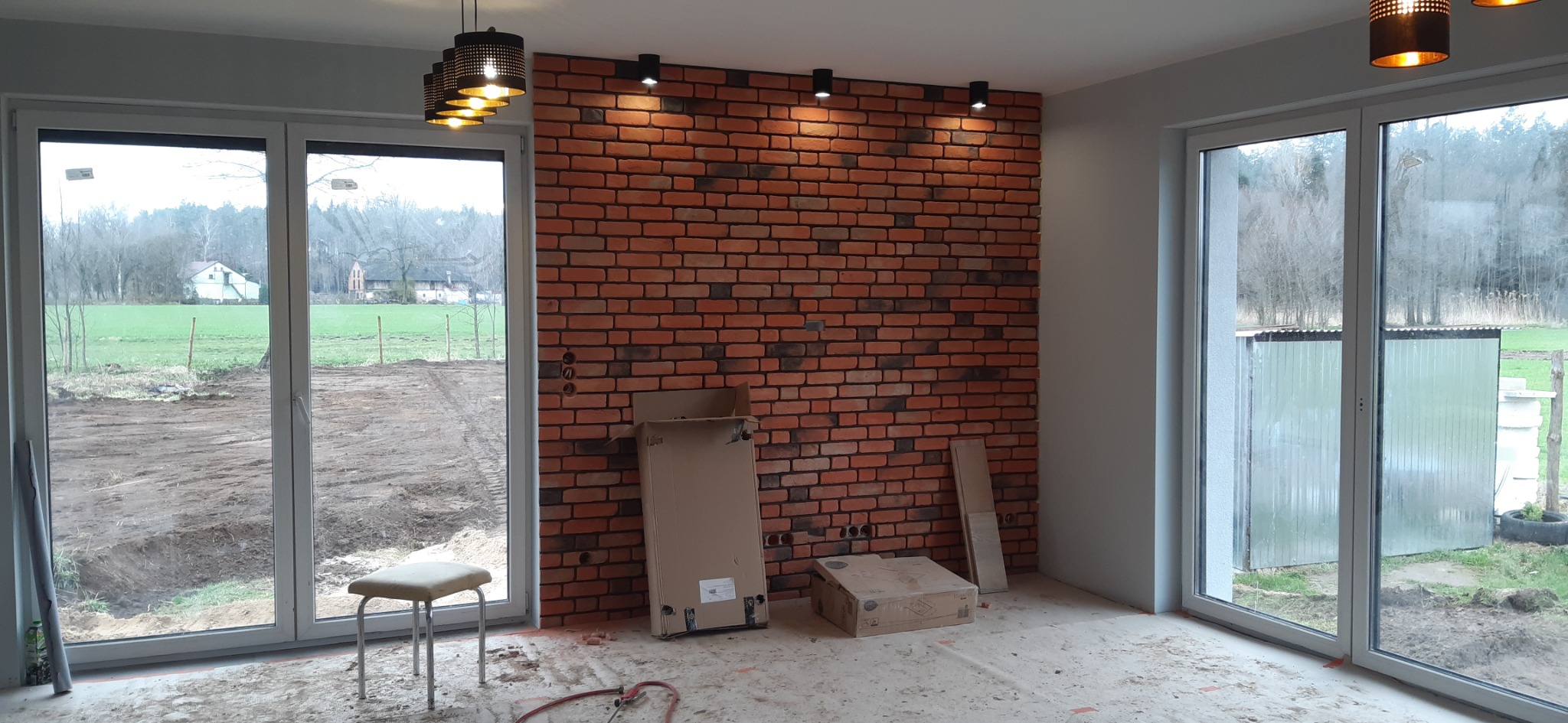Ściana z dekoracyjnej cegły w trakcie montażu, widoczne puszki elektryczne, kartony z materiałami i surowe podłoże, przez okno widok na krajobraz.