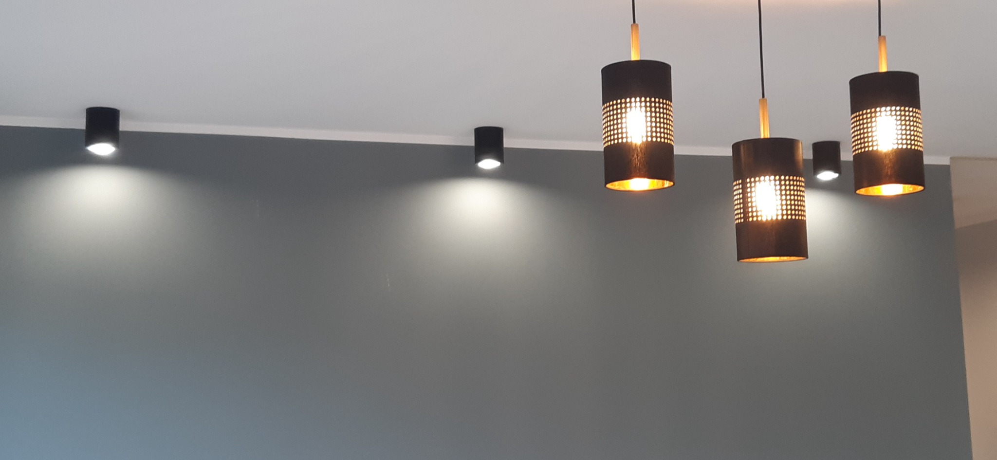 Szara ściana oświetlona przez czarne, cylindryczne lampy sufitowe punktowe oraz wiszące lampy z perforowanym wzorem, dające ciepłe światło.