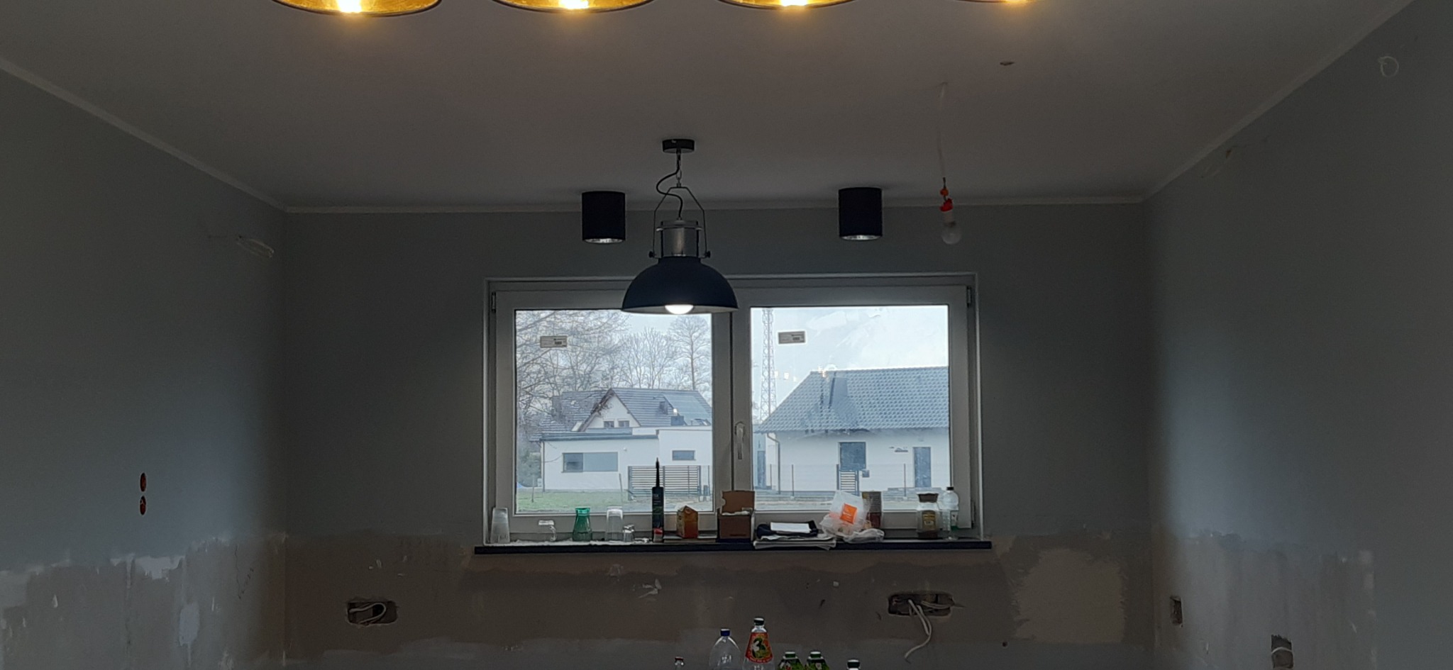 Wnętrze w trakcie przygotowania do malowania: ściany częściowo pokryte szpachlą, widoczne puszki elektryczne, okno z widokiem na domy, sufit z trzema lampami.