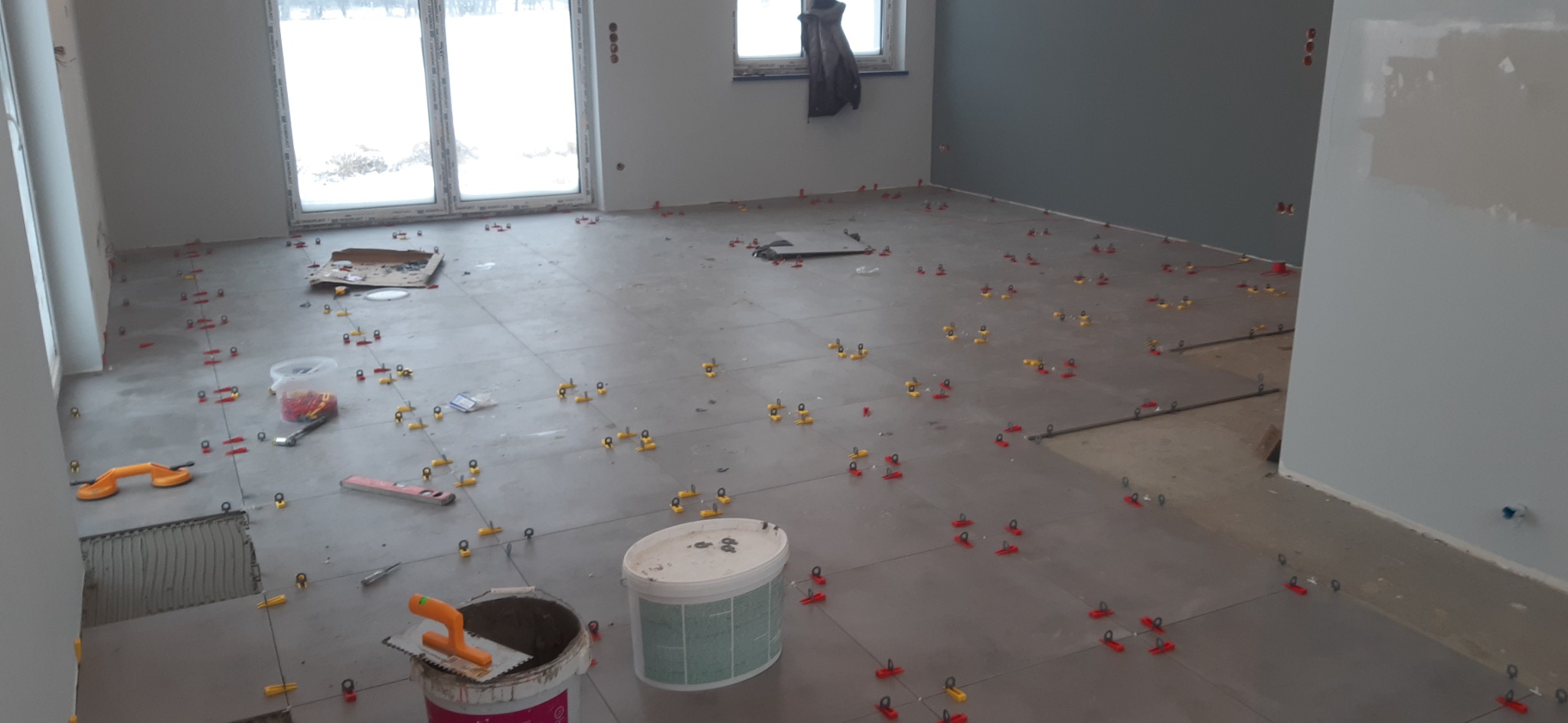 Szare płytki podłogowe w trakcie układania, z widocznymi krzyżykami dystansowymi, przyssawką do płytek i wiaderkami z zaprawą w pomieszczeniu z oknami.
