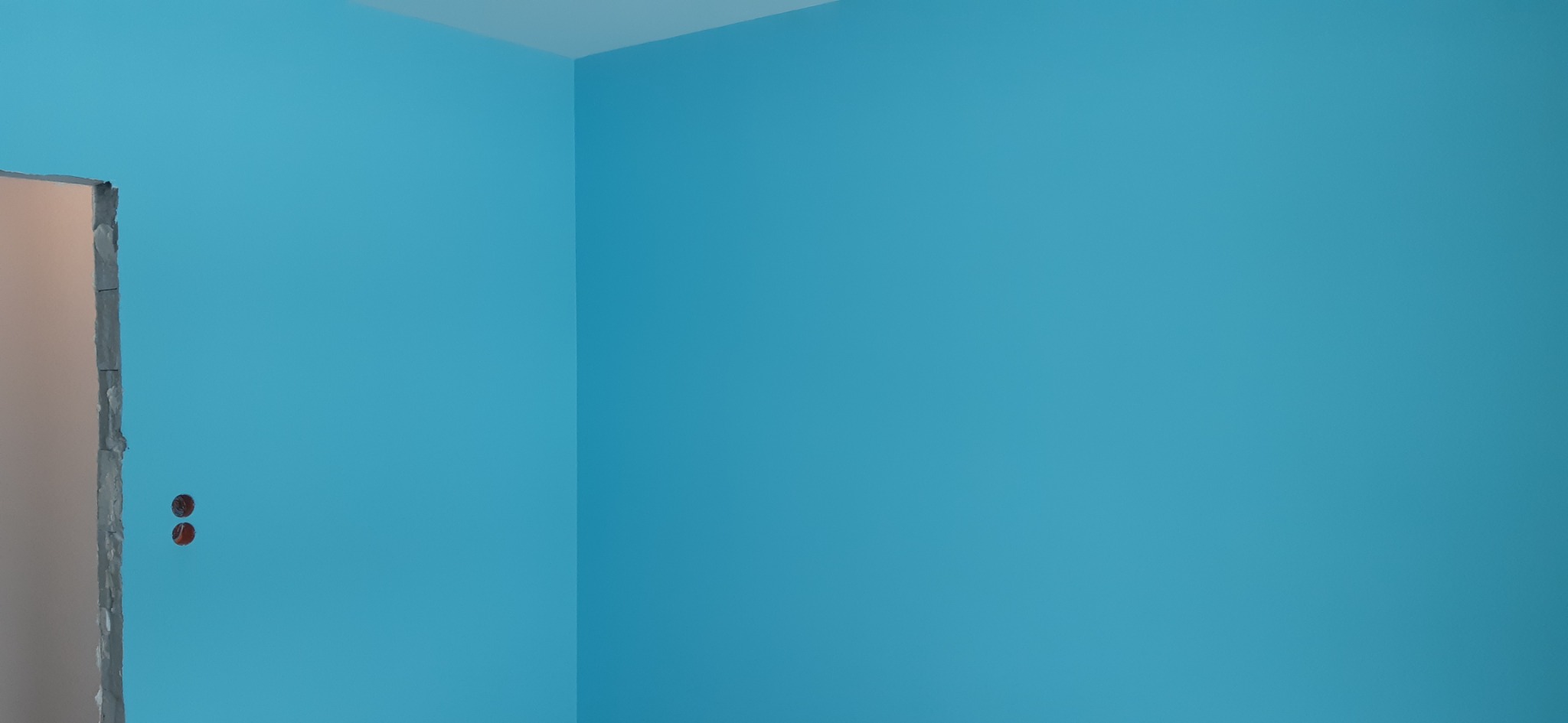 Świeżo pomalowane na błękitno ściany w rogu pokoju, z widocznymi gniazdkami elektrycznymi bez osłon i fragmentem ościeżnicy drzwiowej w trakcie remontu.