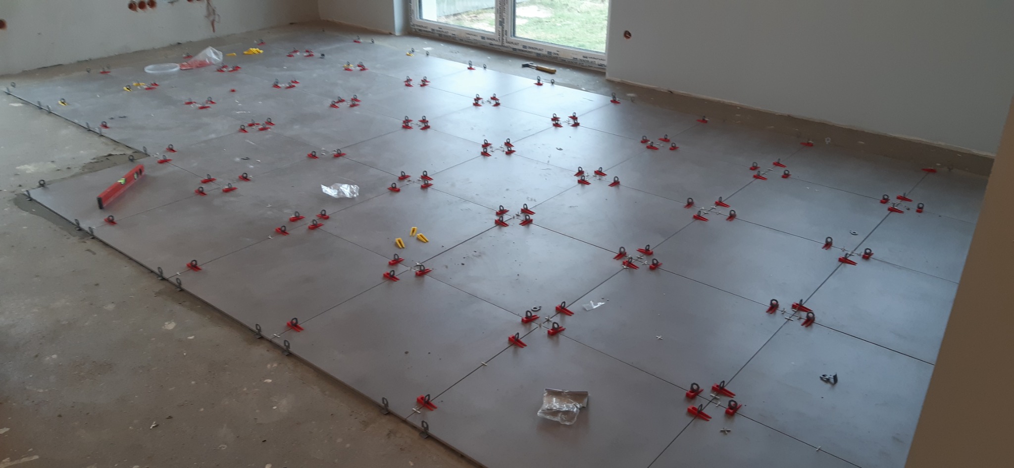 Szare płytki podłogowe w trakcie układania, z czerwonymi i żółtymi klinami dystansowymi, poziomica po lewej stronie, surowy stan ścian i podłogi.