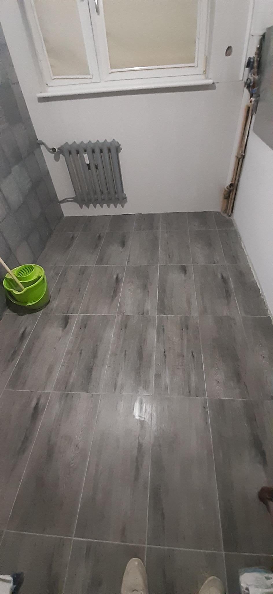 Szare płytki podłogowe imitujące drewno w łazience po remoncie, widoczny grzejnik, okno i wiadro z mopem w rogu.