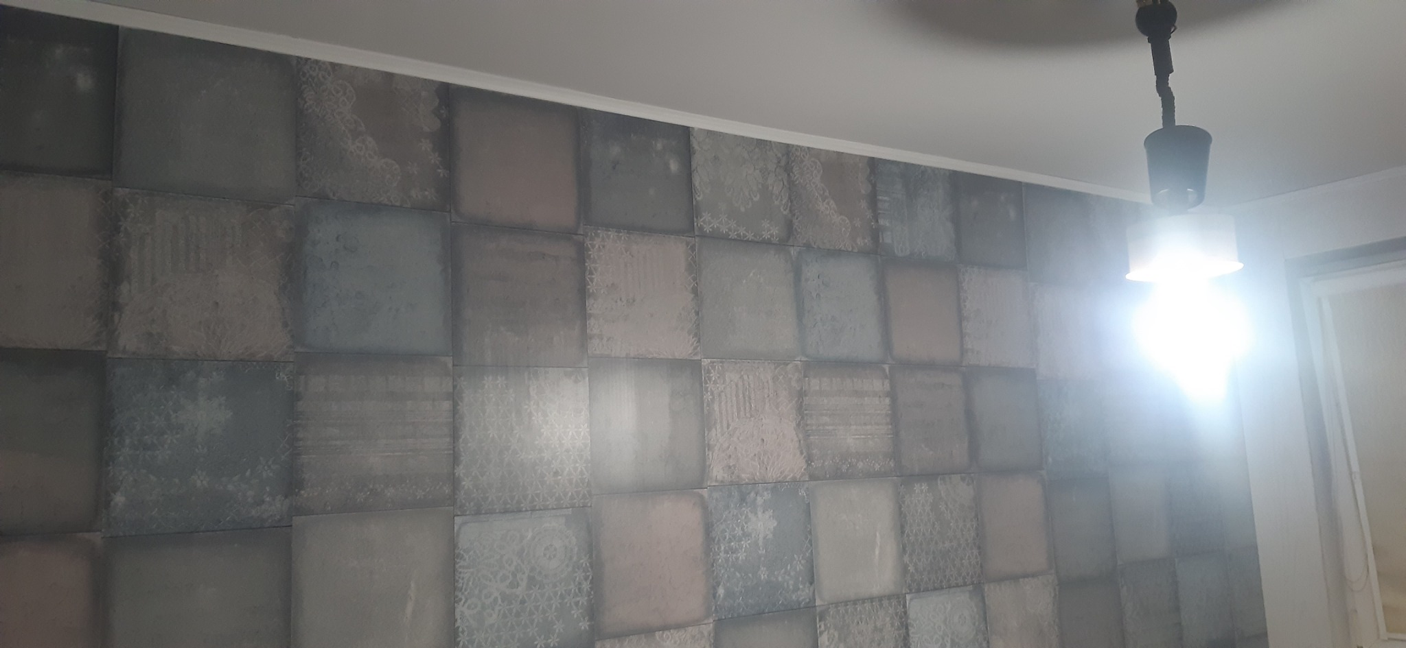 Ściana pokryta tapetą w kwadraty o różnych odcieniach szarości i wzorach, oświetlona przez lampę sufitową z widocznym kablem.