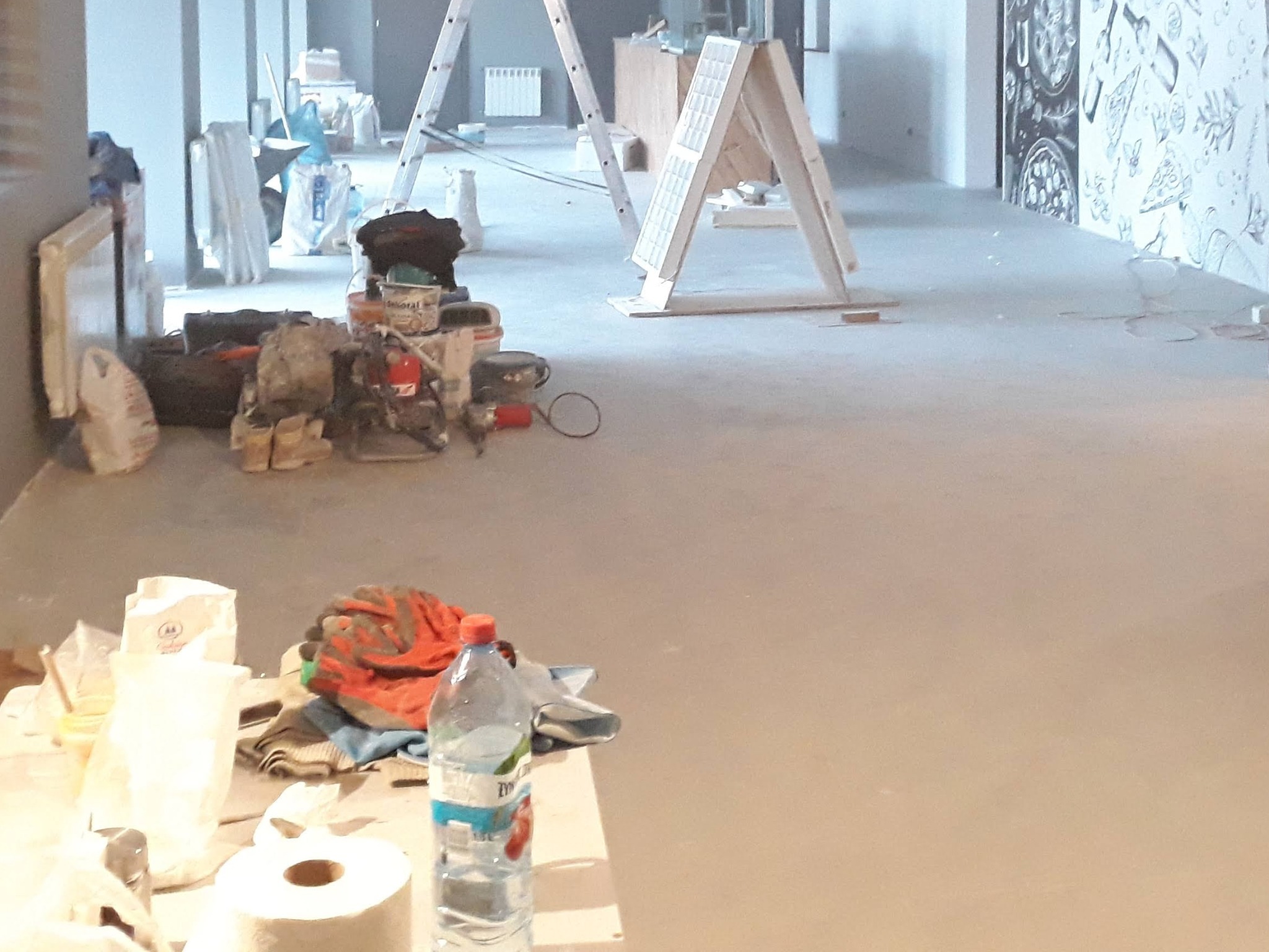 Wnętrze w trakcie prac wykończeniowych, widoczne narzędzia, drabina, puszki z farbą, worki z materiałami budowlanymi, zabezpieczona ściana z dekoracyjnym malunkiem.
