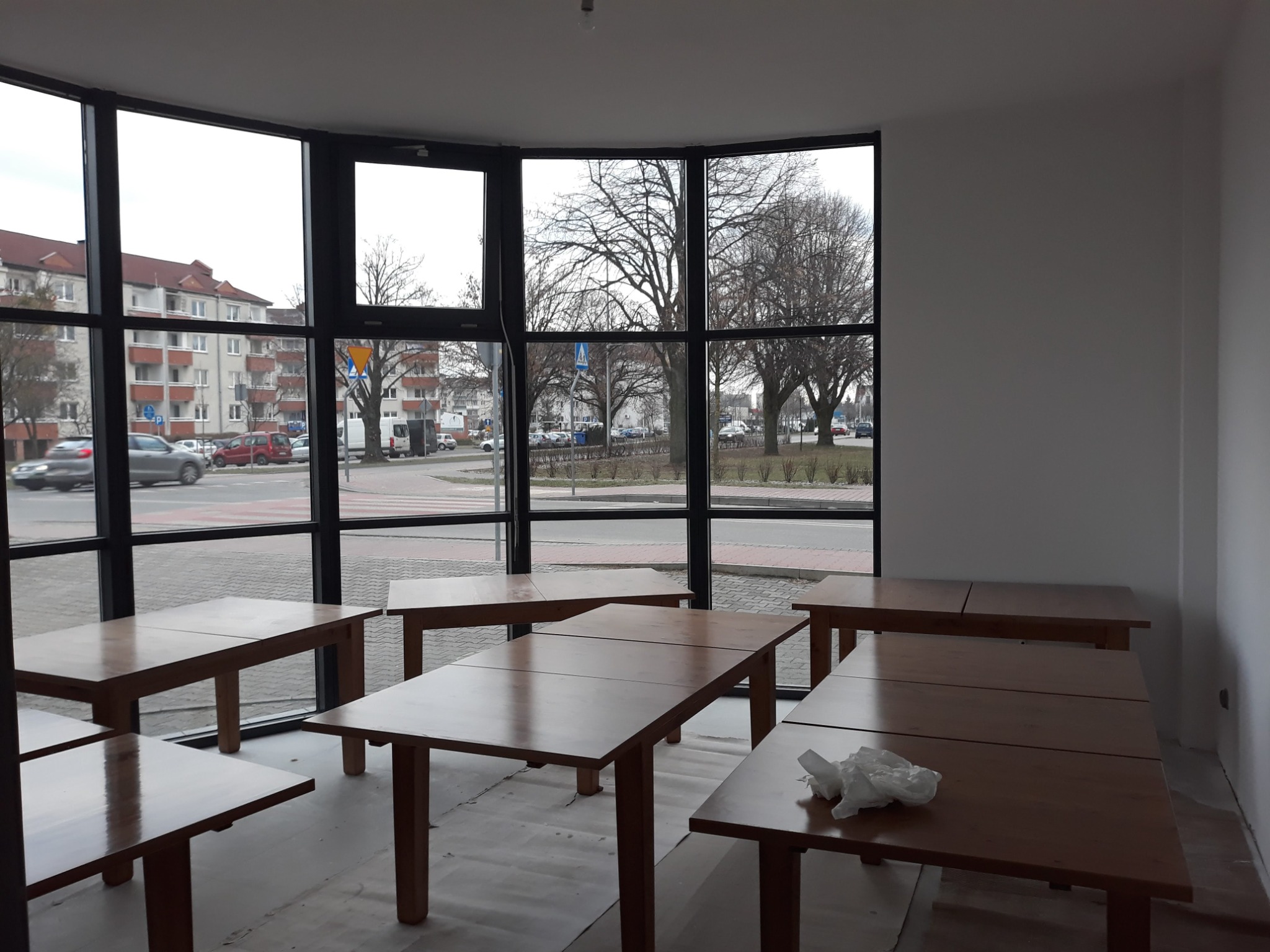 Jasne wnętrze biura z dużymi oknami i drewnianymi stołami, widoczne niedoskonałości wykończenia przy podłodze i narożniku, sugerujące etap przygotowania do użytku.