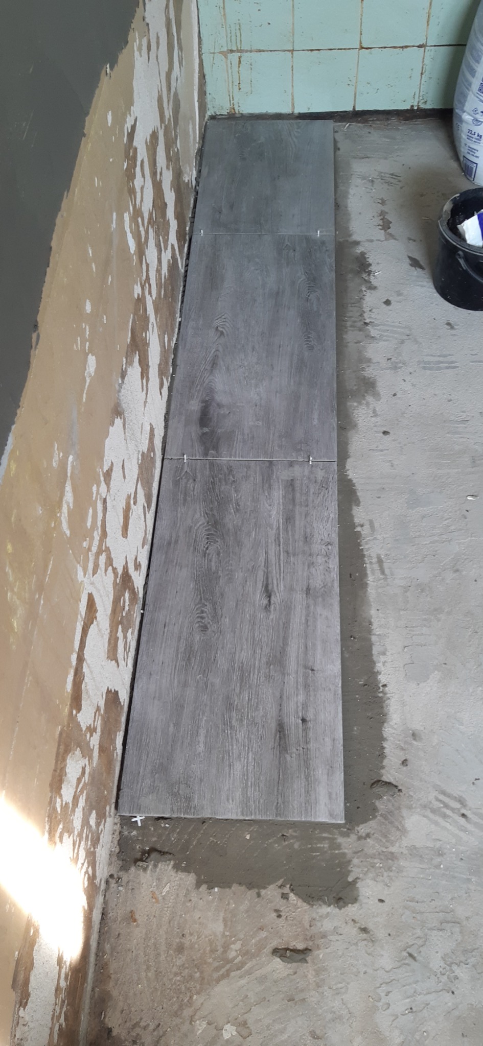Układanie szarych płytek podłogowych imitujących drewno, widoczne krzyżyki dystansowe, surowa ściana w tle z resztkami starej farby.