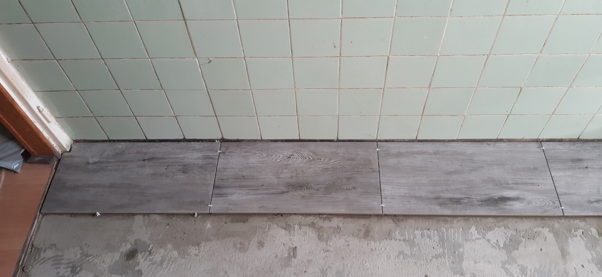 Fragment podłogi w trakcie kładzenia płytek imitujących drewno, widoczne krzyżyki dystansowe, stare, kwadratowe płytki na ścianie i surowy beton podłoża.