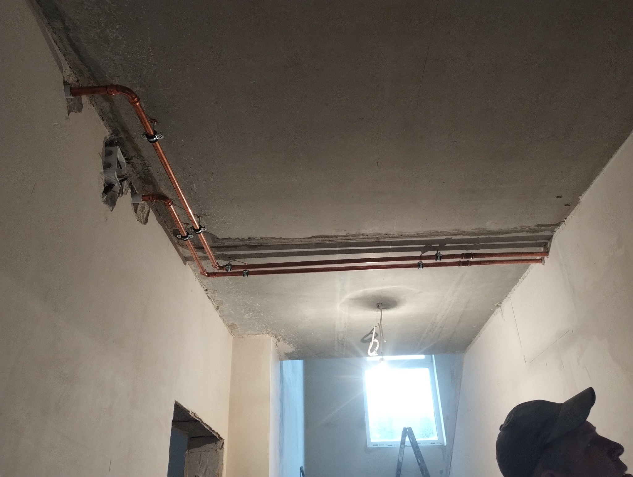 Miedziane rury centralnego ogrzewania zamontowane pod betonowym sufitem podczas remontu, widoczne mocowania i fragment instalacji elektrycznej w ścianie.